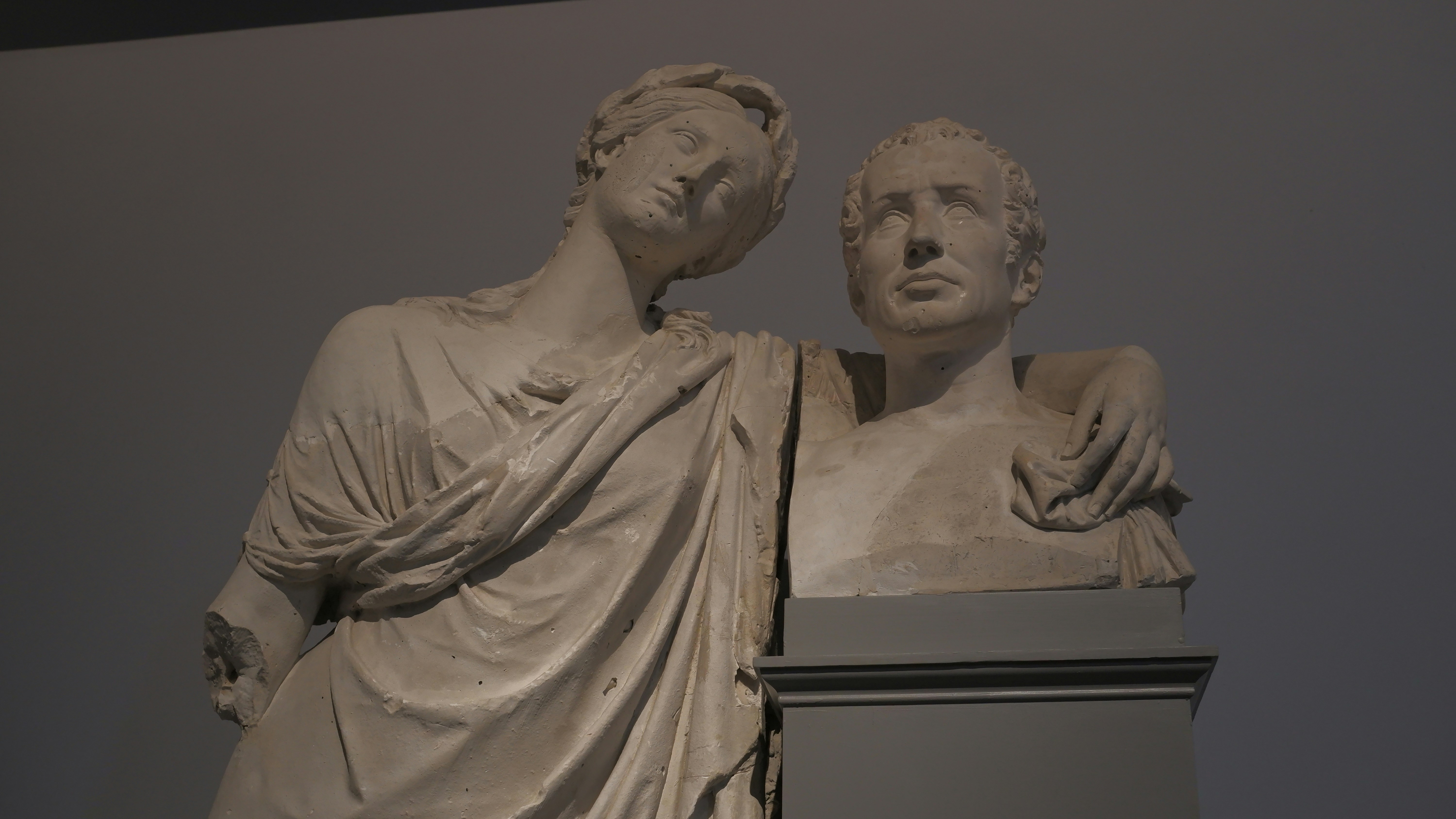Two classical marble busts embrace on pedestal.