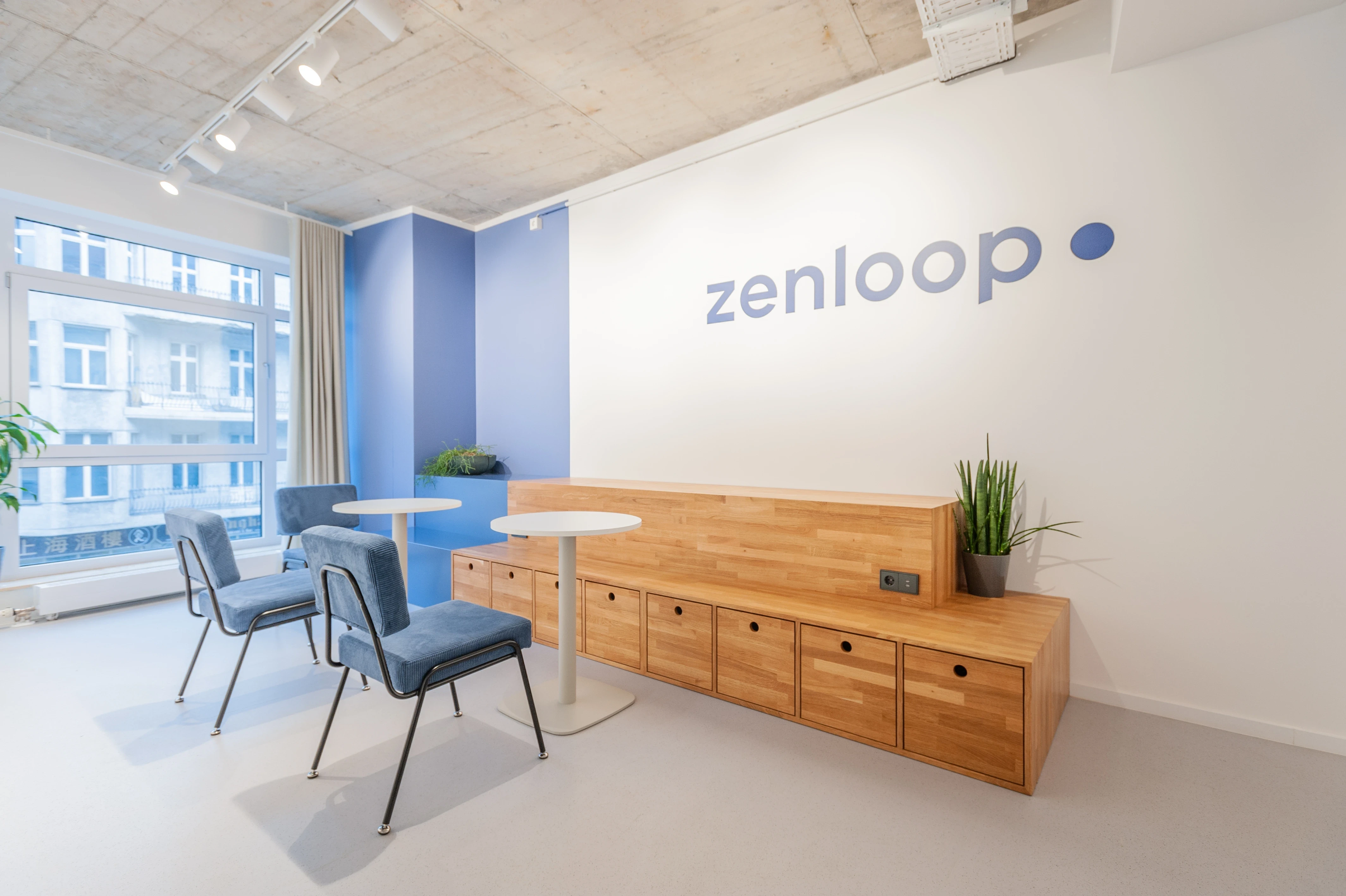 Reception lounge at ZENLOOP with a pink desk in front of a wooden slat wall with navy logo, a cream sofa and black chair by large windows and exposed concrete ceiling.