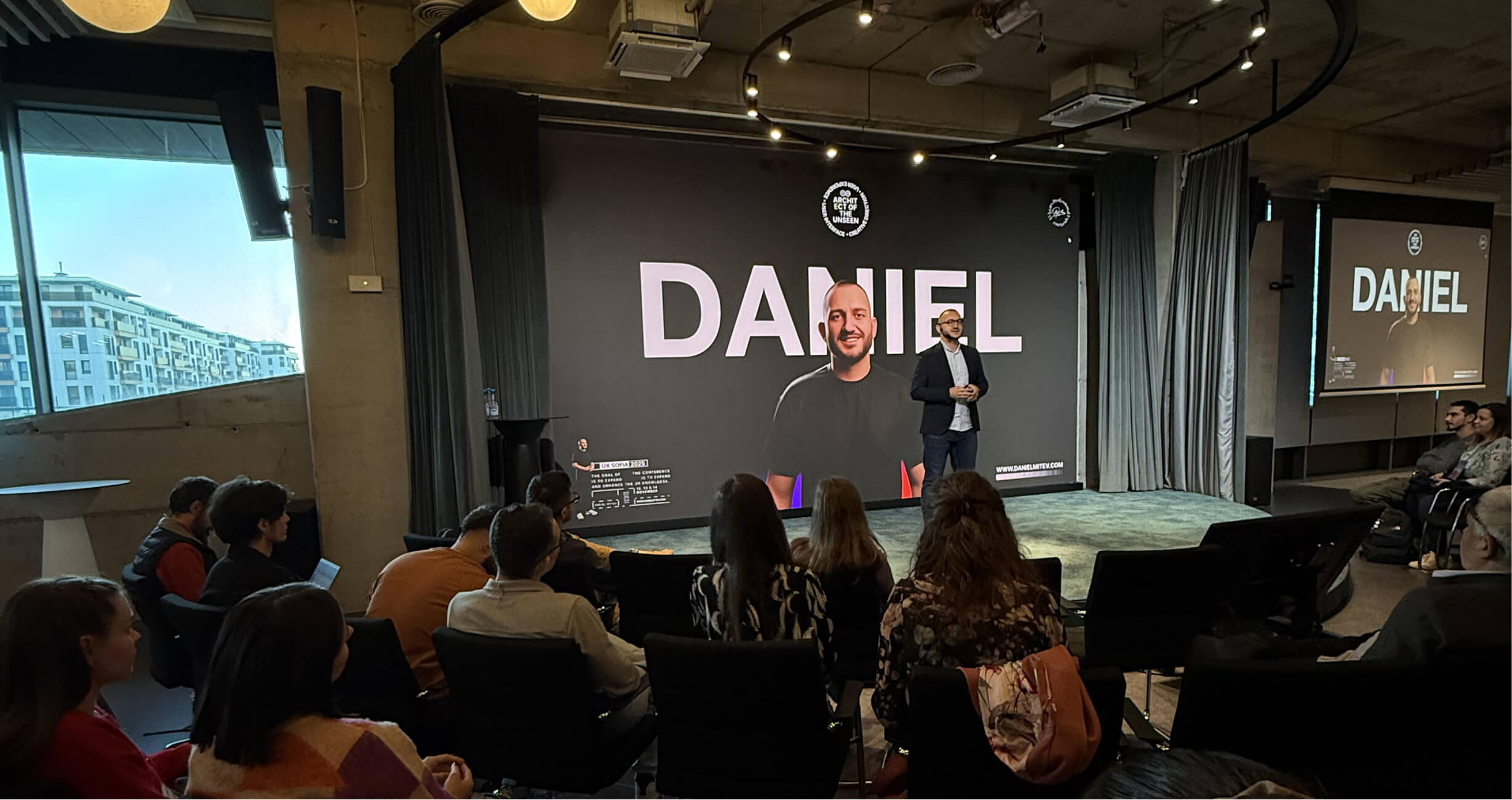 A speaker presents on a stage with a large image and text that reads "DANIEL" projected behind them, in a modern conference room filled with seated attendees.