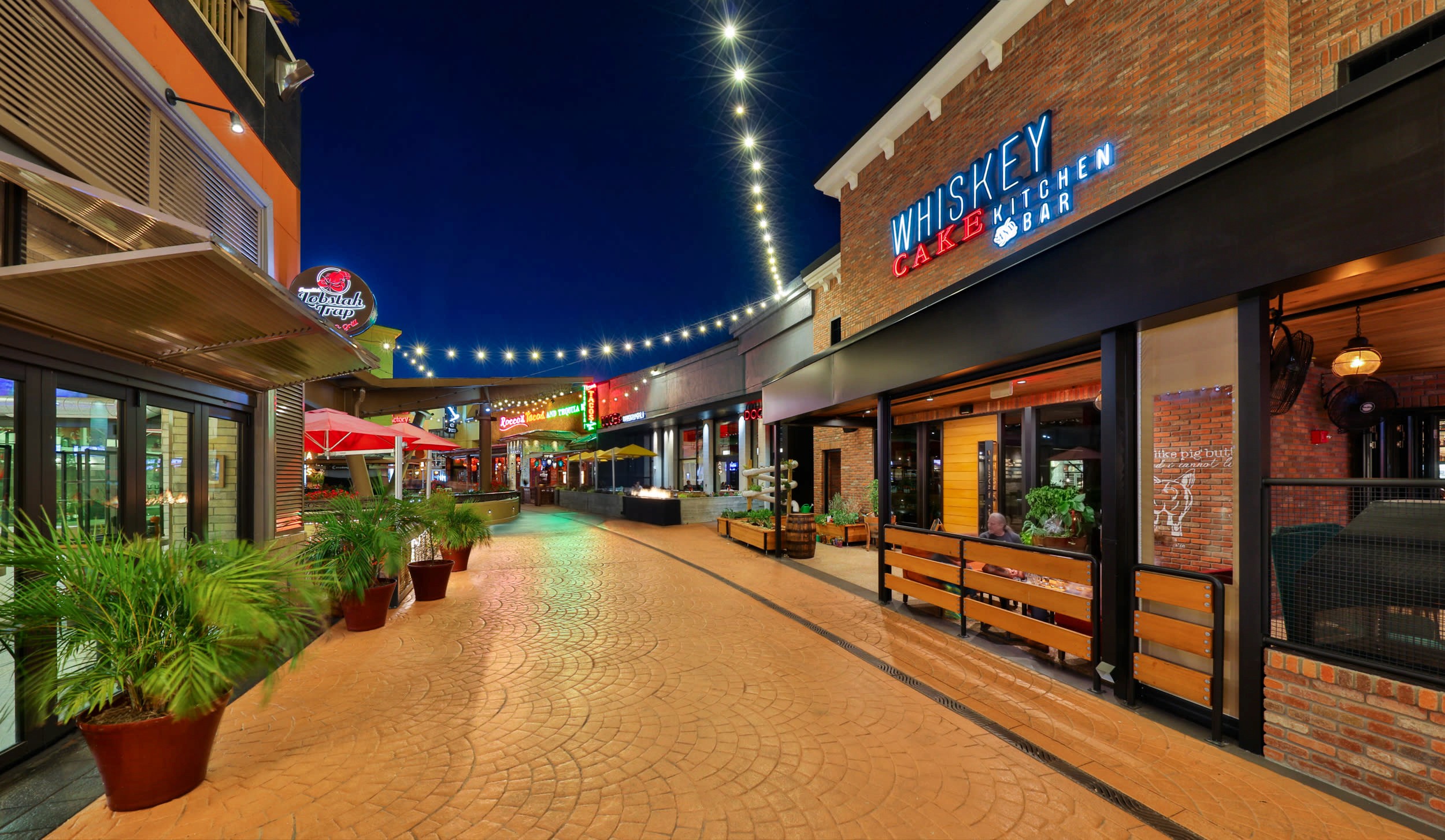 Rendering of International Plaza's expansion showing a modern open-air shopping street with storefronts, warm lighting, landscaping, and pedestrian walkways in the evening
