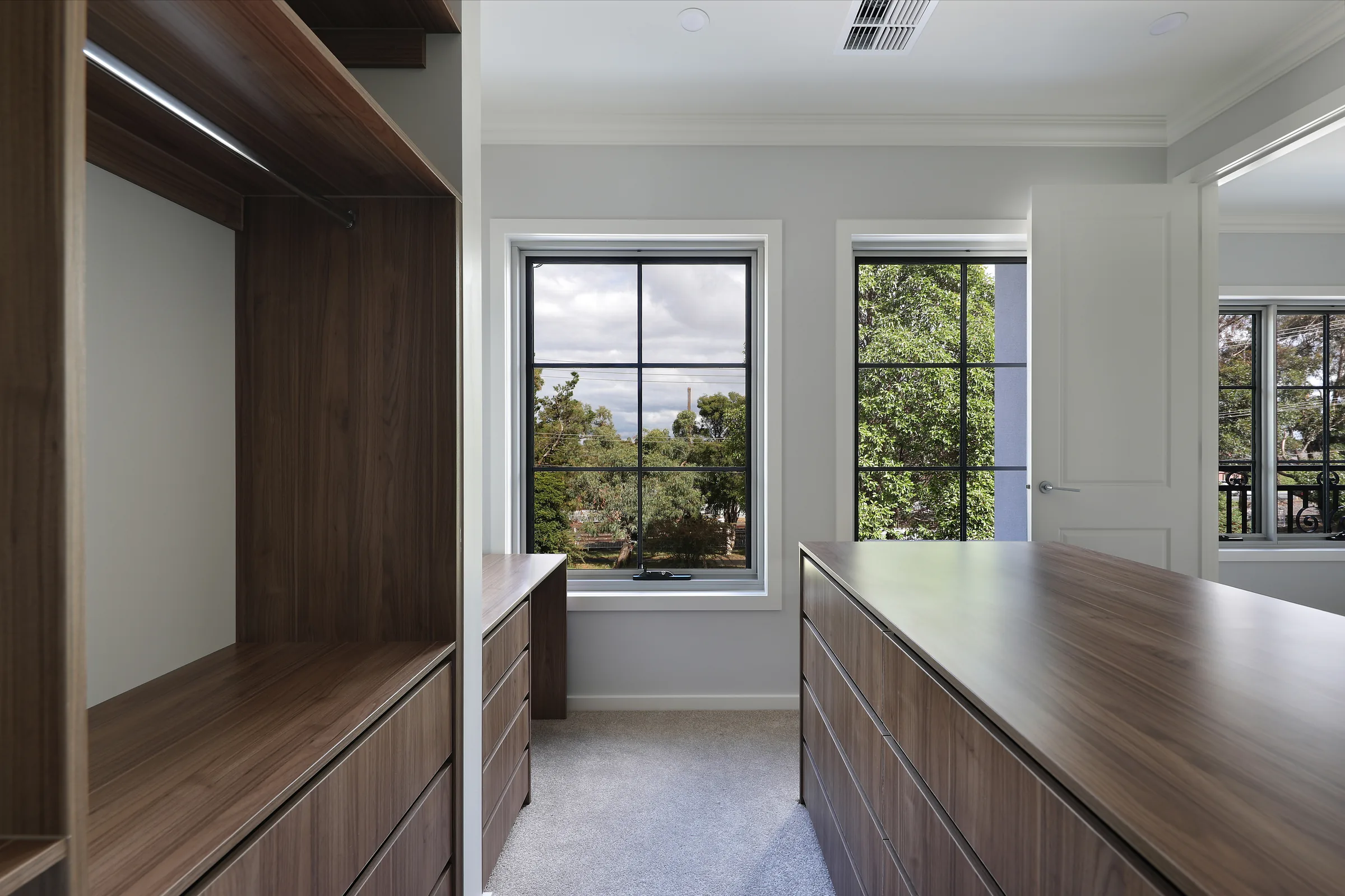 Walk-in dressing room of the Glen Waverley Residence with walnut-lined joinery, open hanging rails, banks of drawers, grey carpet and black-framed double-hung windows looking onto leafy trees
