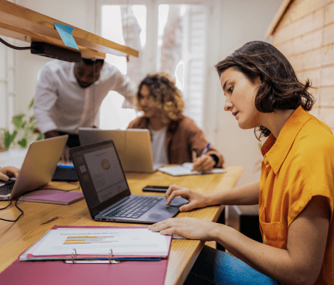 A focused woman in an orange shirt is working on a laptop at a table surrounded by colleagues. She appears to be reviewing documents and data on her screen. In the background, two other people are engaged in discussion, with laptops and notebooks on the table, indicating a collaborative work 