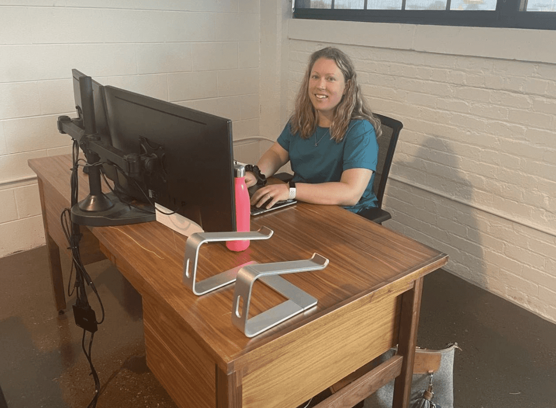 A person sitting at a desk with a large monitor, looking towards the camera, in a well-lit room.