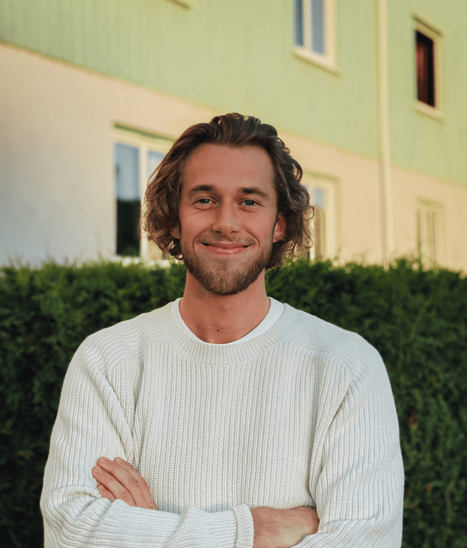 A portait of Lukas, he have a big smile, short beard and long blond hair. Wearing a knitted sweater outside.