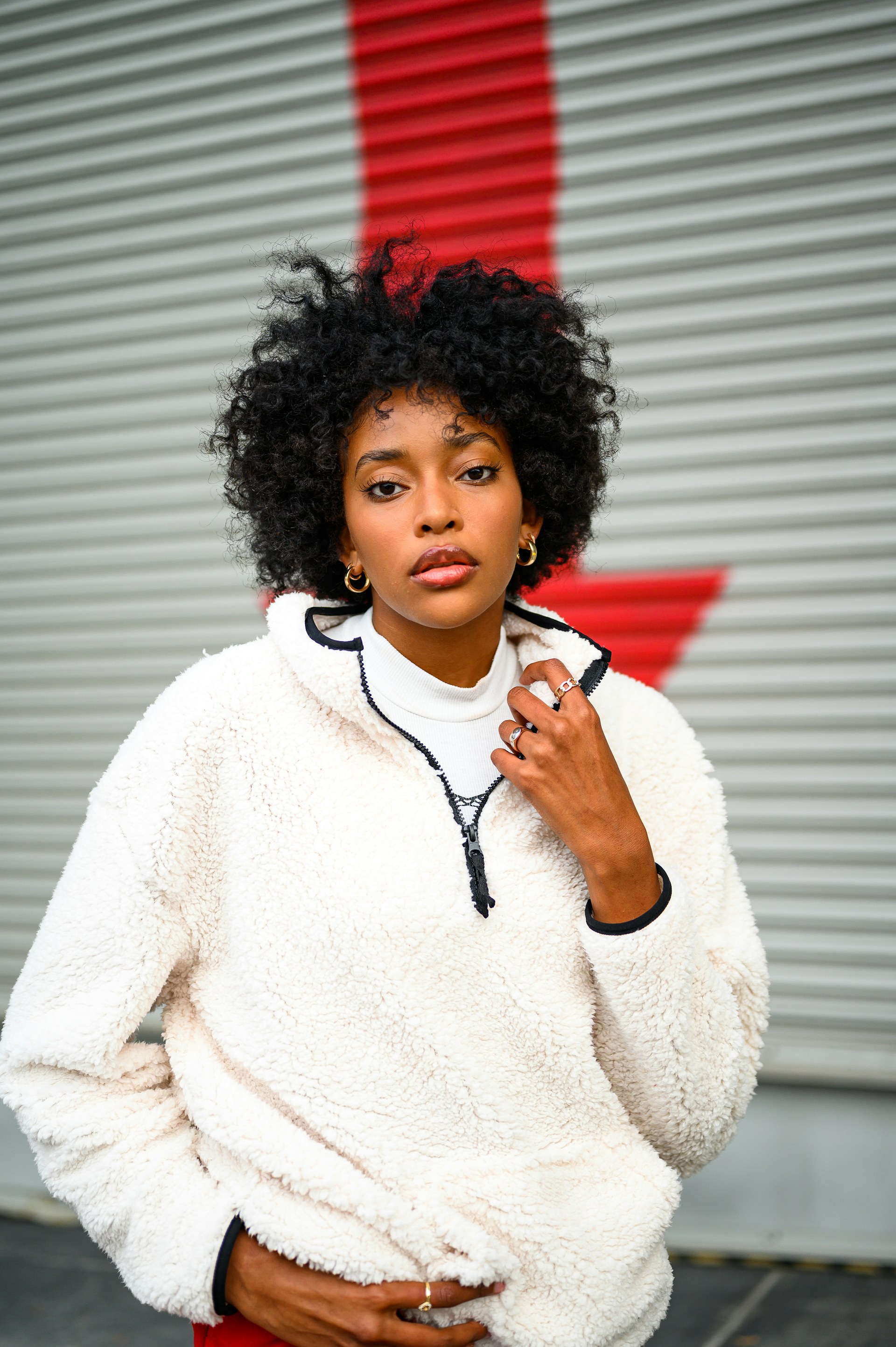 A woman with a natural afro and gold hoop earrings looking directly at the camera. She is wearing a white sherpa quarter-zip fleece over a white turtleneck, set against a grey and red arrow background.
