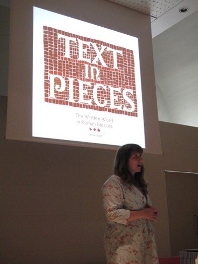 A photo of a woman giving a lecture while having behind her the projected white text on a red brick background that says: Text in Pieces.