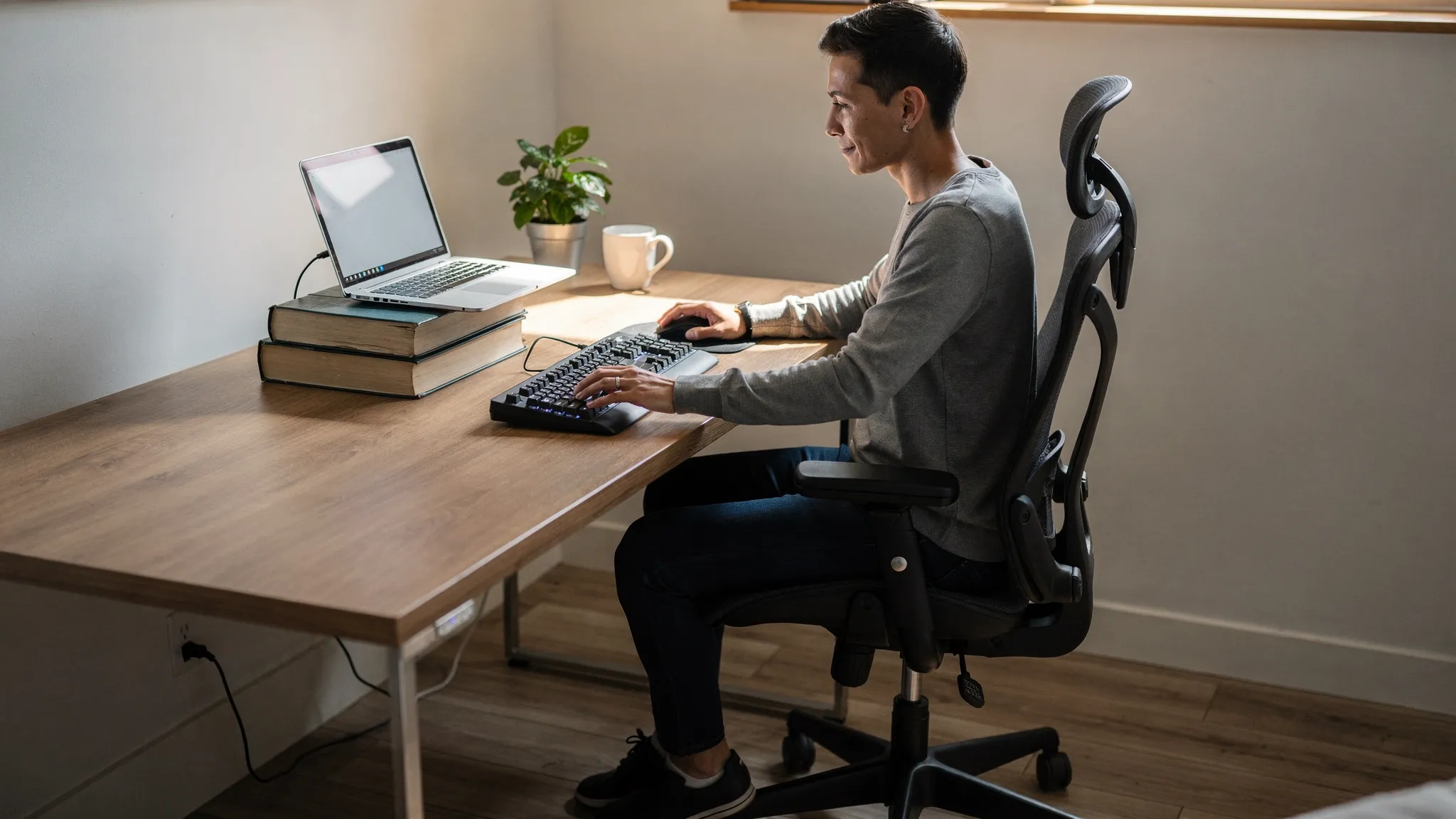 Person at a home office desk adjusting their chair height and moving the keyboard closer, with a laptop raised on books and a separate keyboard and mouse visible.