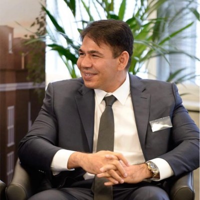 Smiling man with a beard and styled hair, wearing a black suit and white shirt, photographed against a plain gray background. Professional and friendly tone.