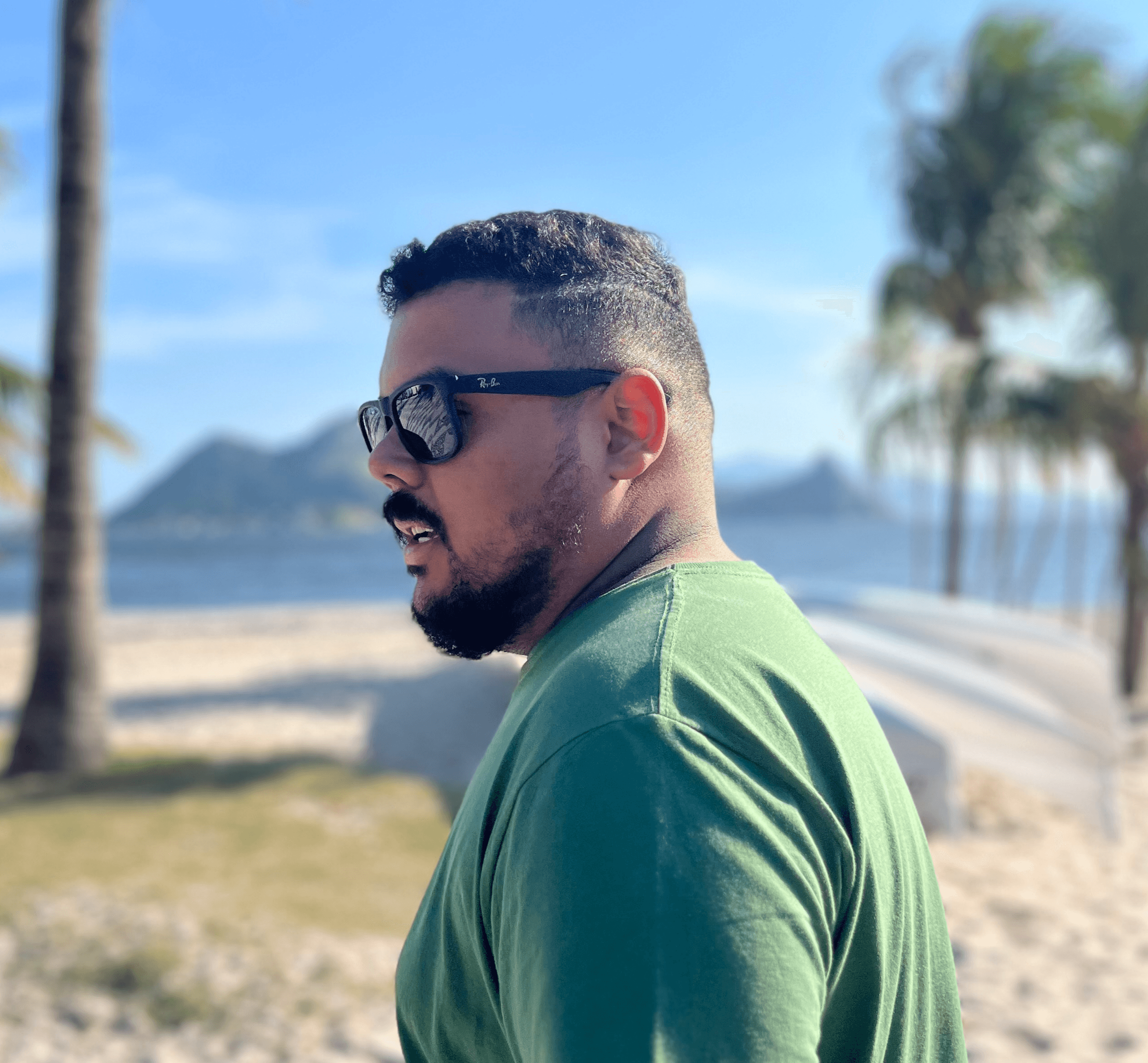 A man with sunglasses and a beard, wearing a green shirt, standing near a beach with palm trees and the ocean in the background.