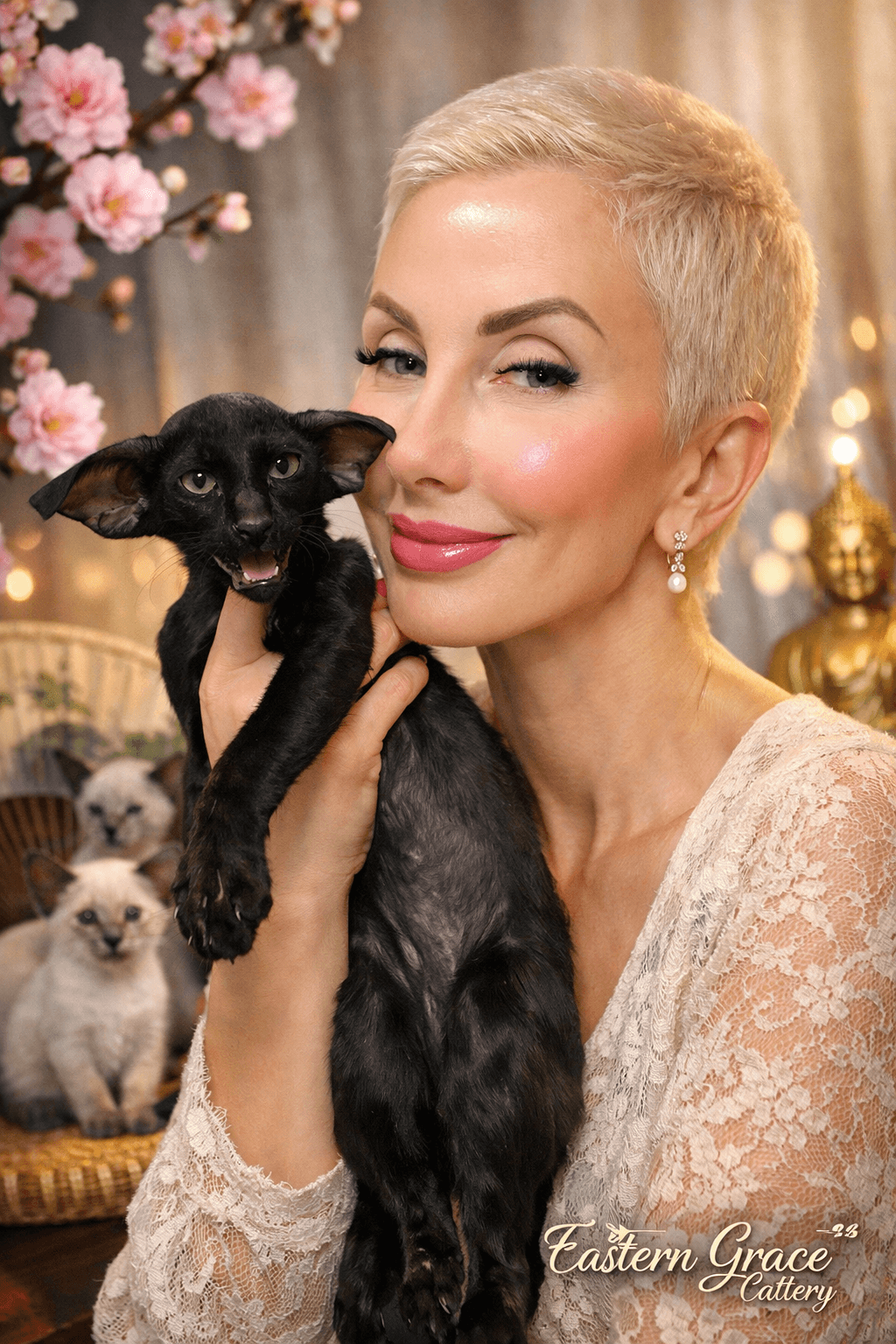 A smiling person with short blonde hair gently holds a black Oriental Shorthair cat in a cozy indoor setting with Siamese kittens in the background and decorative pink blossoms, creating an elegant and serene atmosphere.
