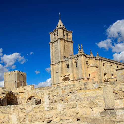 Isang makasaysayang simbahang gawa sa bato na may mataas na kampanaryo, napapalibutan ng mga guho sa ilalim ng maliwanag na asul na langit na may kaunting ulap.