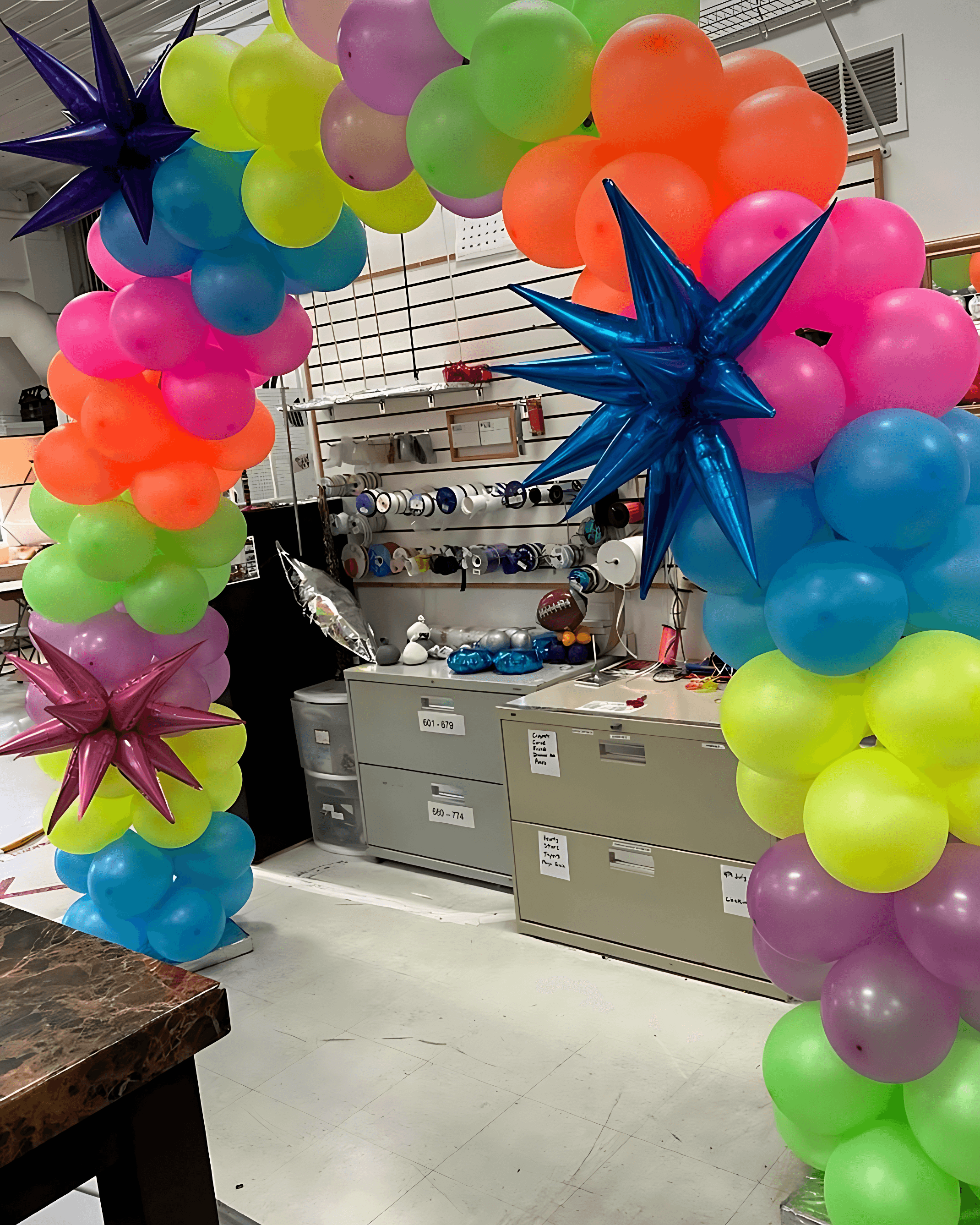 A vibrant balloon arch in a workshop features a mix of colorful balloons in neon shades of pink, orange, yellow, green, and blue, adorned with shiny star-shaped balloons, set against a background of tools and supplies.