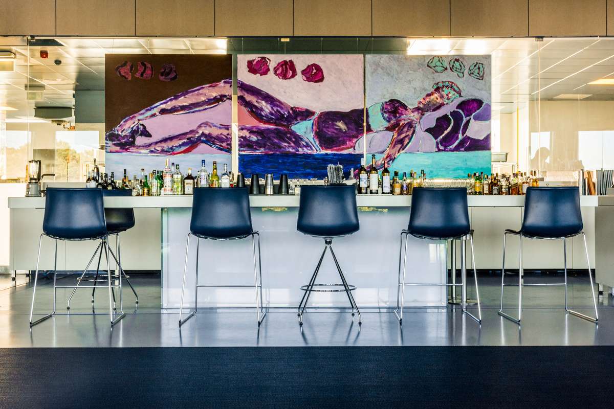 Five modern blue chairs sit in front of a long white counter beneath a large abstract painting with purple and pink tones.