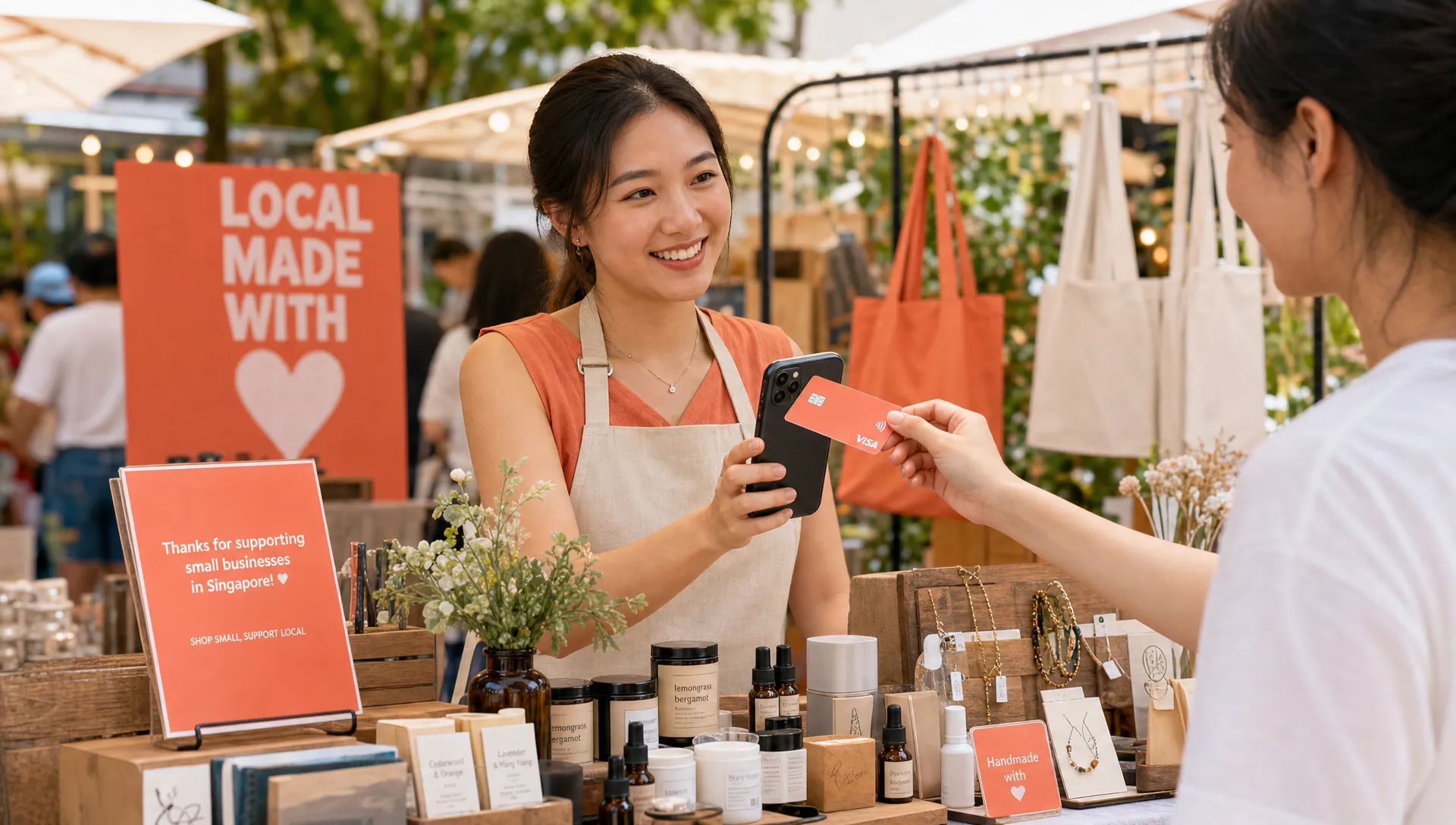 A small Singapore business owner accepting a contactless card payment on a smartphone at a market-style pop-up booth, with neatly arranged products on the table and a customer tapping a card on the back of the phone.