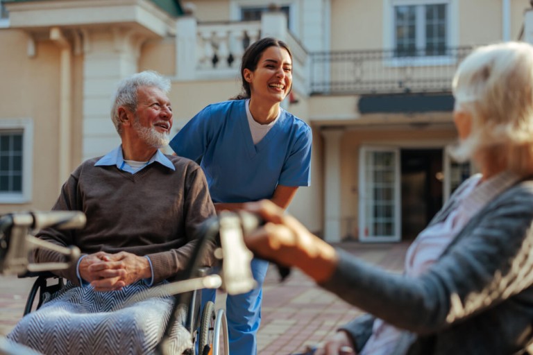 A nurse in blue scrubs smiling while assisting an elderly man in a wheelchair outdoors in front of a building