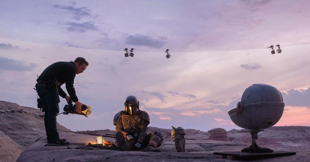 Two people work outdoors at dusk, one holding a light source while the other sits nearby, surrounded by equipment.