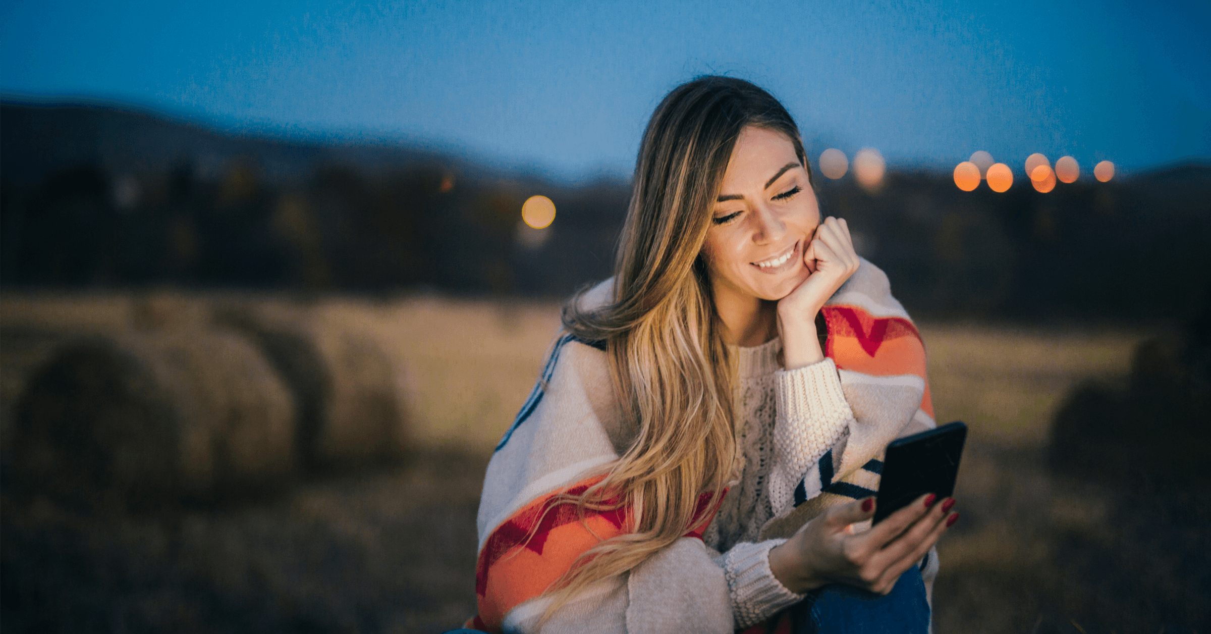 En kvinna sitter och ler till sin mobil ensam utomhus på kvällen.