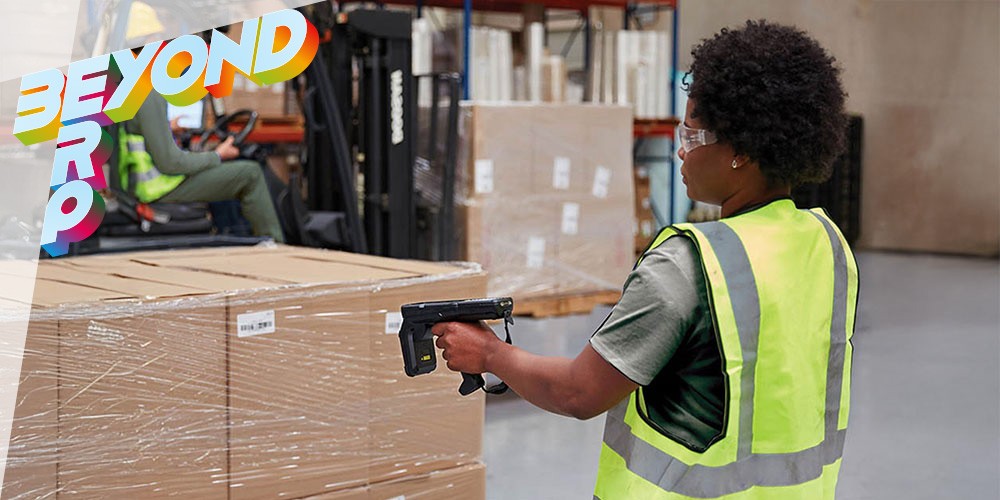 A person in a safety vest holds a scanner while looking at a pallet of boxes and flowers in a warehouse.