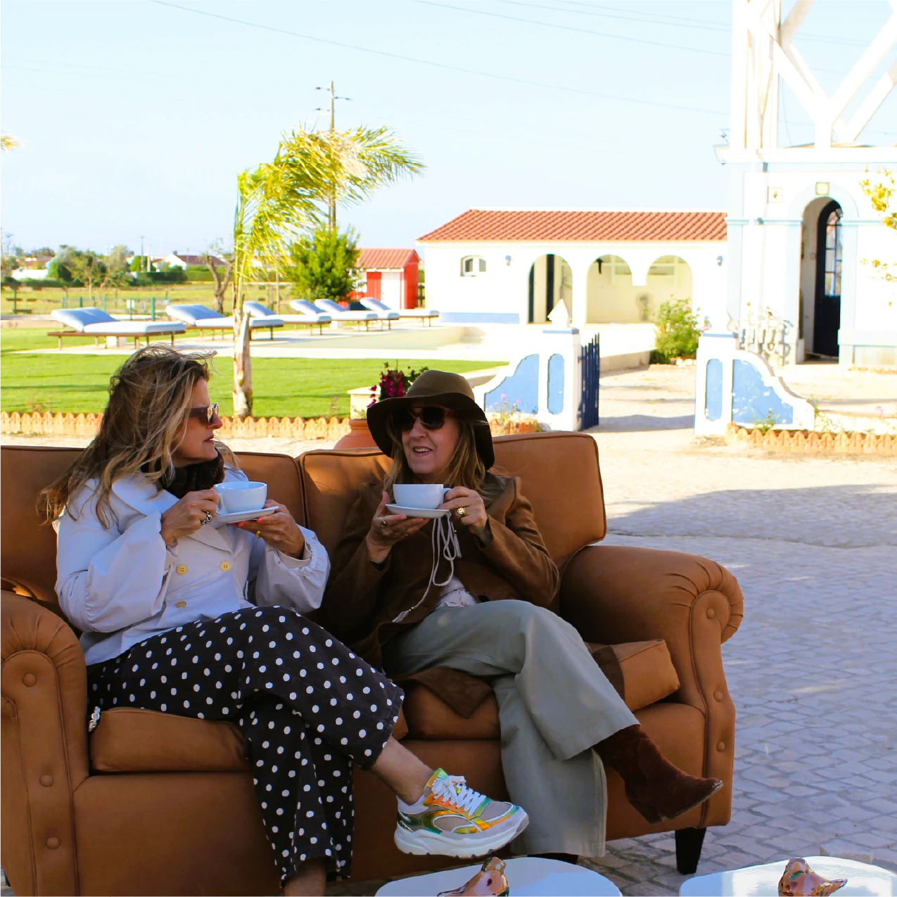 Two women sit on an outdoor sofa, sipping tea. They are dressed casually and appear relaxed, enjoying a sunny day in a garden setting.