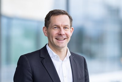 A smiling man in a dark suit stands in front of a blurred background, suggesting a professional setting.