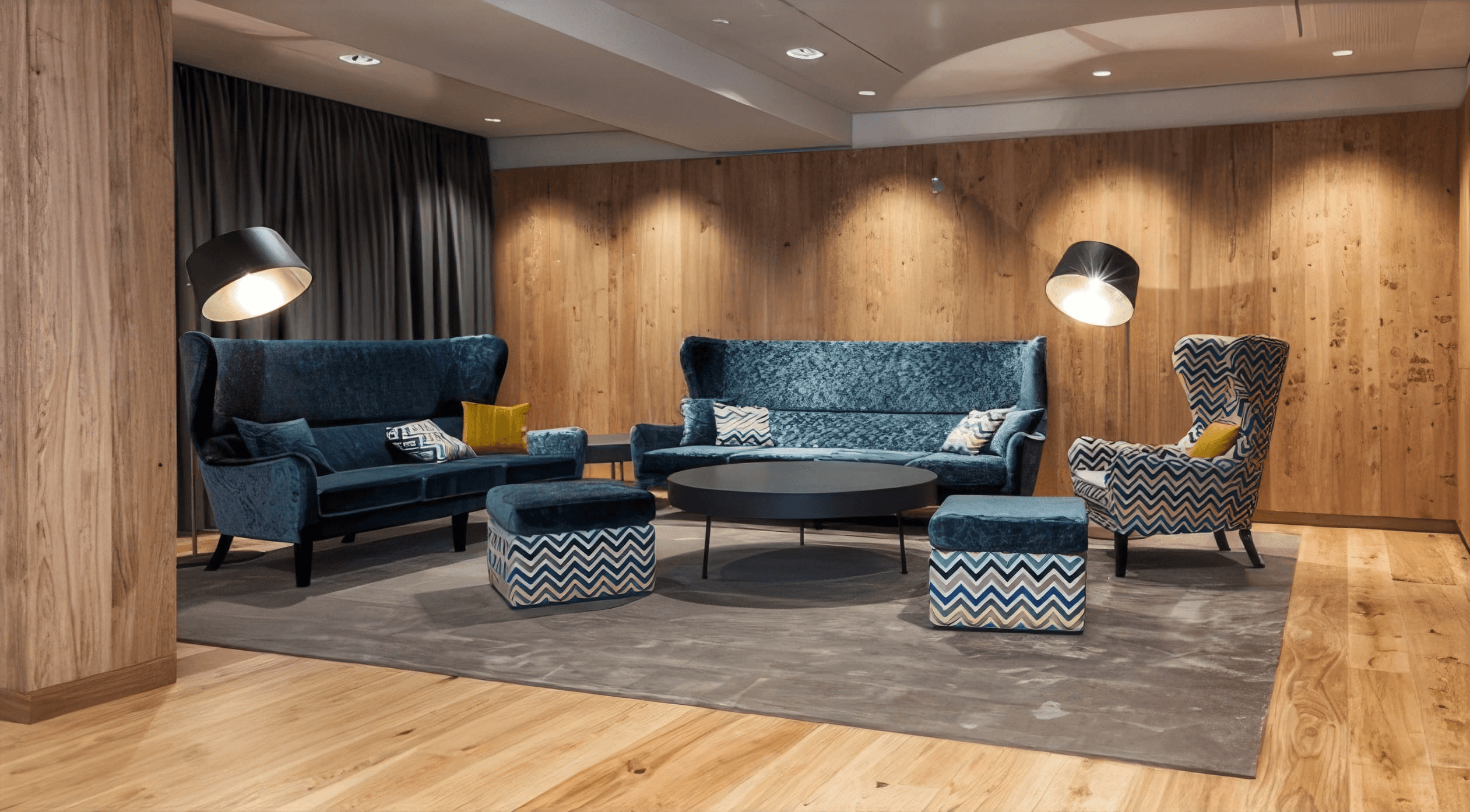 MW Interior  - Gemütlicher Hotellobby-Loungebereich mit blauen Samt-Sofas, gemusterten Hockern, rundem Couchtisch und warmer Holzwandverkleidung" Beschreibung: Einladende Hotellobby mit tiefen blauen Samtsofas, gemusterten Zickzack-Hockern in Blau-Gold, schwarzem rundem Couchtisch und warmer Eichenholzwand – ein Wohnzimmergefühl mit alpin-elegantem Charakter.