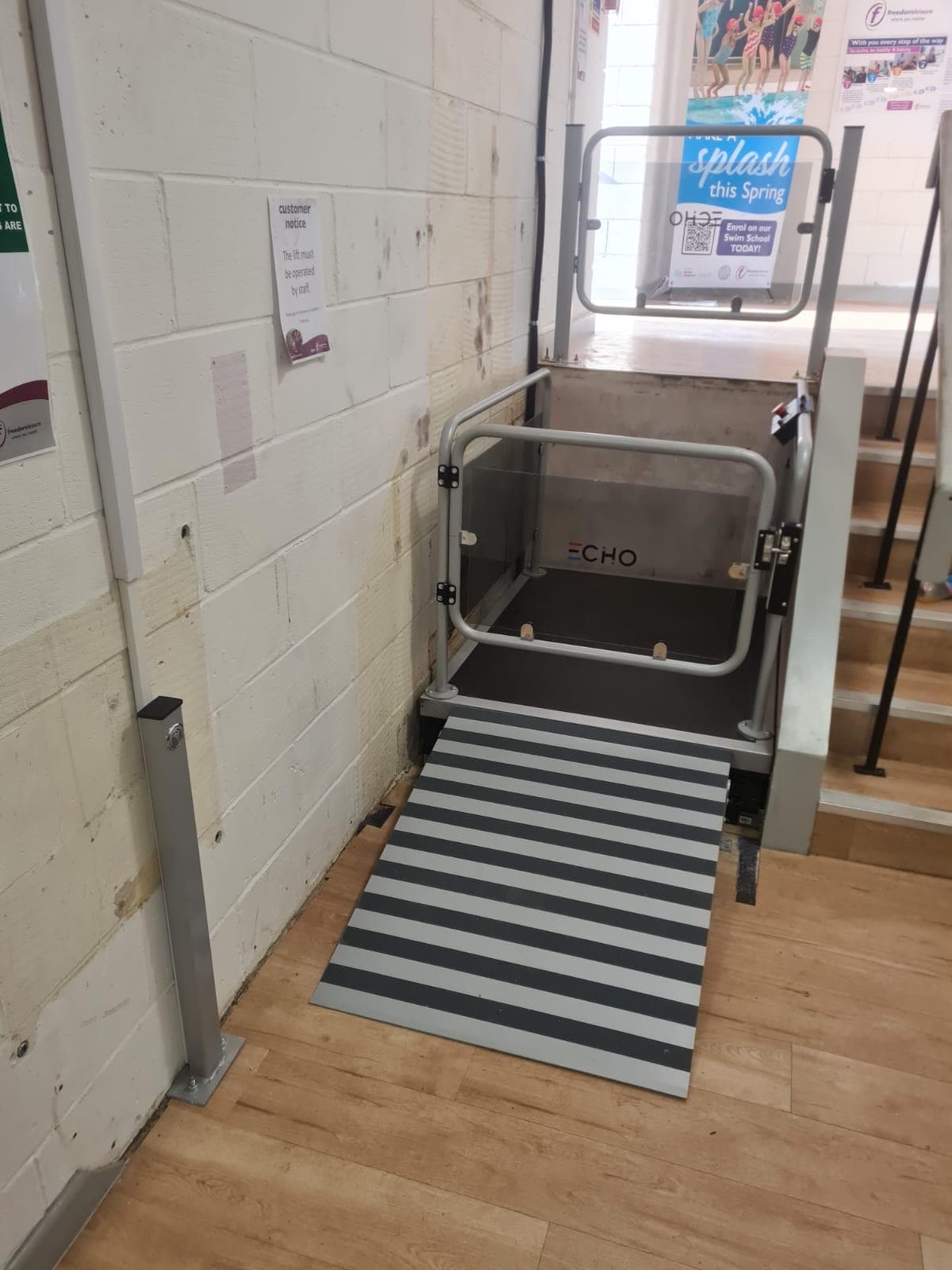 Echo commercial vertical platform step lift at lower landing level inside a UK leisure centre — grey tubular safety frame with tinted glass panels and striped anti-slip entry ramp deployed