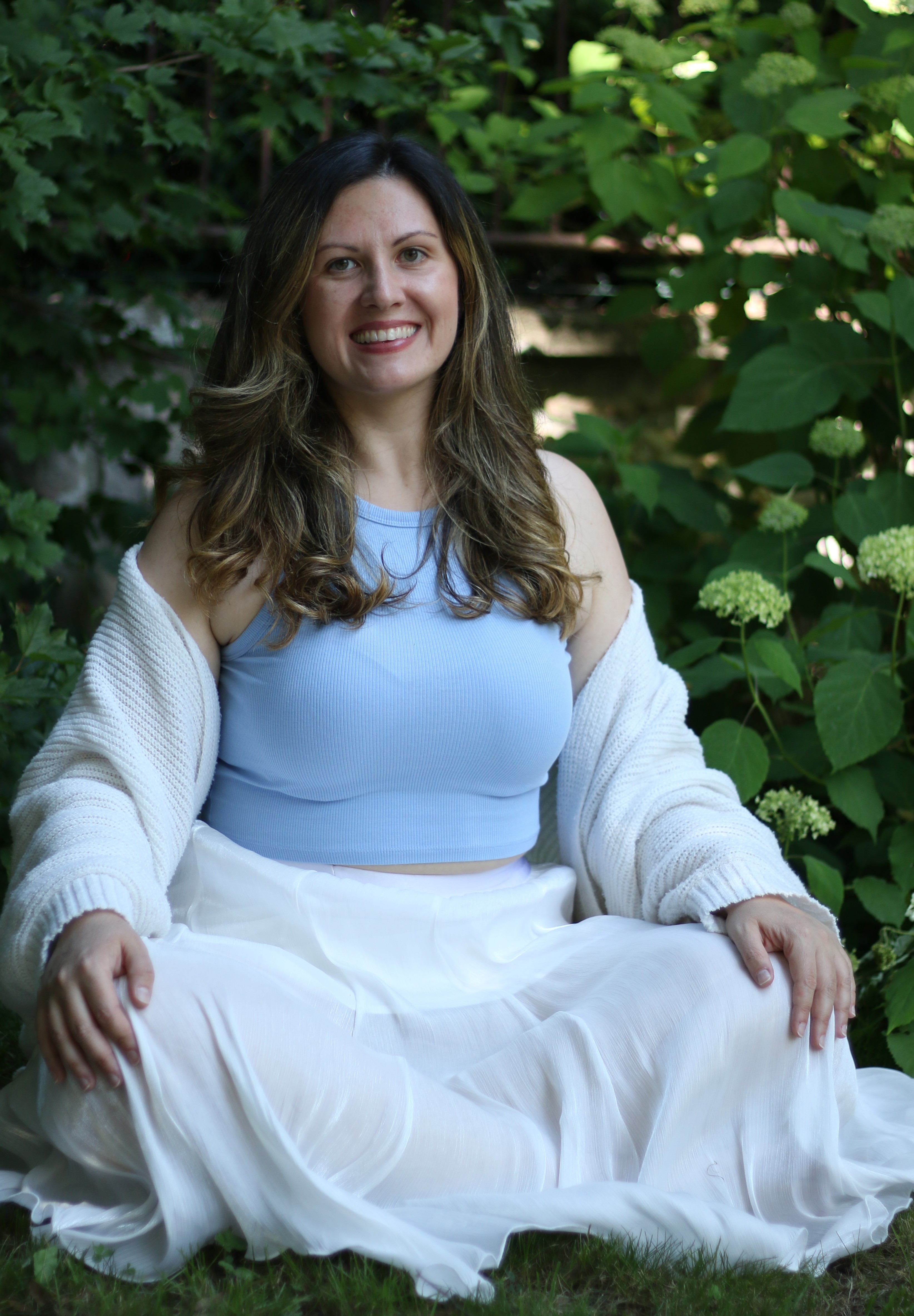 Woman sitting cross-legged outdoors, smiling, wearing a light blue top and white cardigan.
