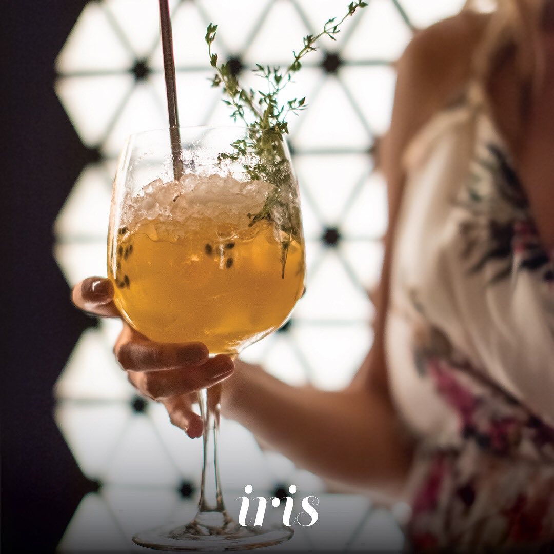 A hand holding a garnished cocktail in a wine glass, with patterned screen in the background, IRIS logo in the bottom.