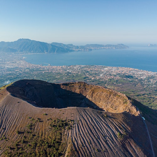 Aerial view of a volcanic crater with surrounding ridged terrain, overlooking a coastal city and mountainous landscape.