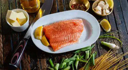 Salmon fillet on a white plate with lemon wedges, surrounded by butter, garlic, pepper, green onions, pasta, and a knife on a rustic wooden table.