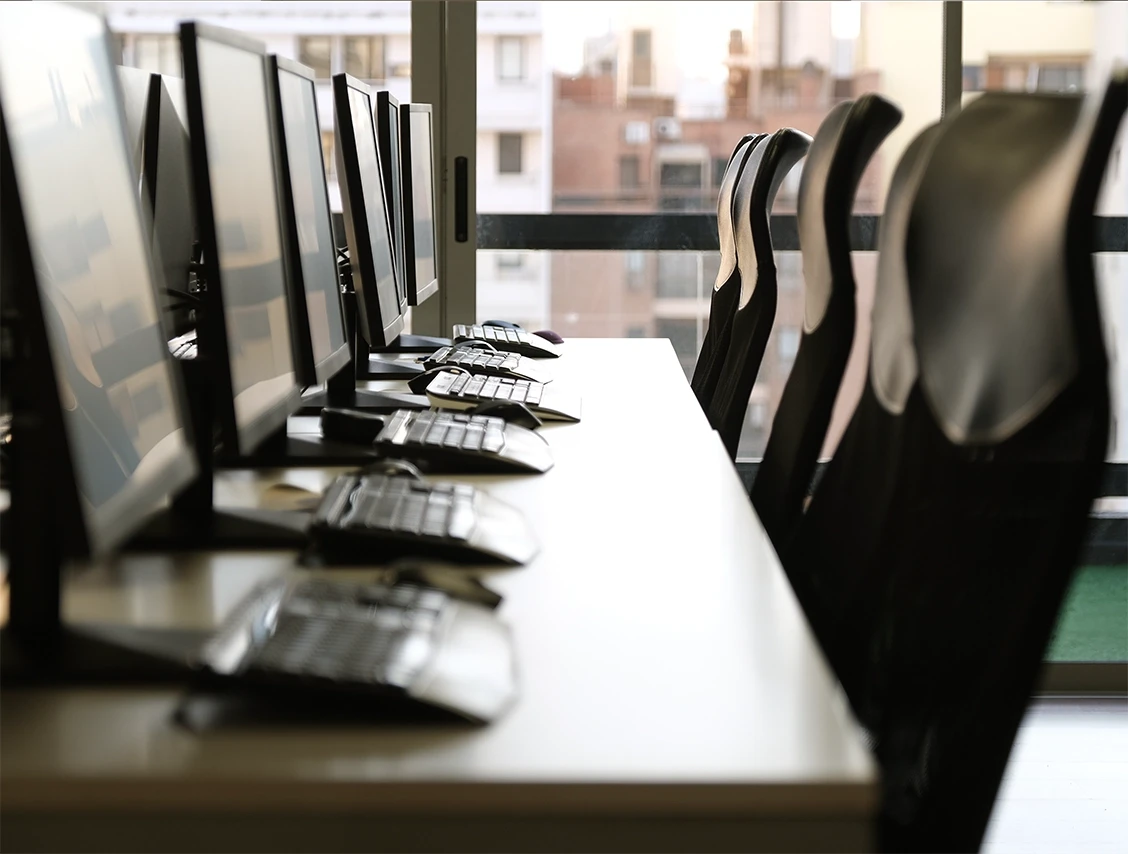 Side view of aligned computer monitors and ergonomic chairs in a bright, glass-walled office.