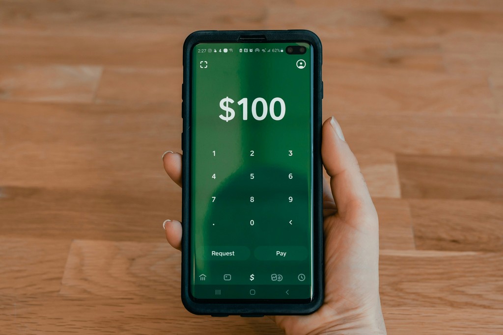 A hand holding a smartphone with a finance app open, displaying a transaction of $100 on the screen. The app features a simple and modern design with large numbers, set against a green background. The wooden table beneath and the warm tone of the hand provide a natural contrast to the device's technology.