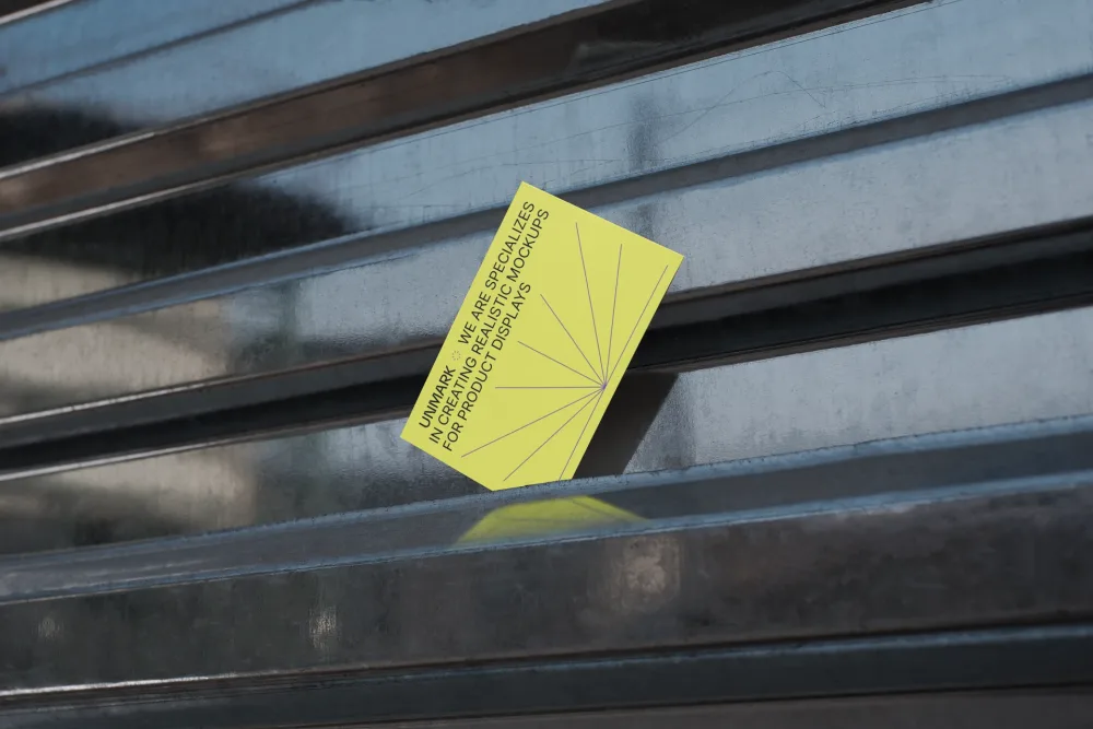 Bright yellow card leaning against a reflective metal surface, featuring diagonal line accents and minimal text. Sunlight creates shadows and subtle reflections on the metallic background.