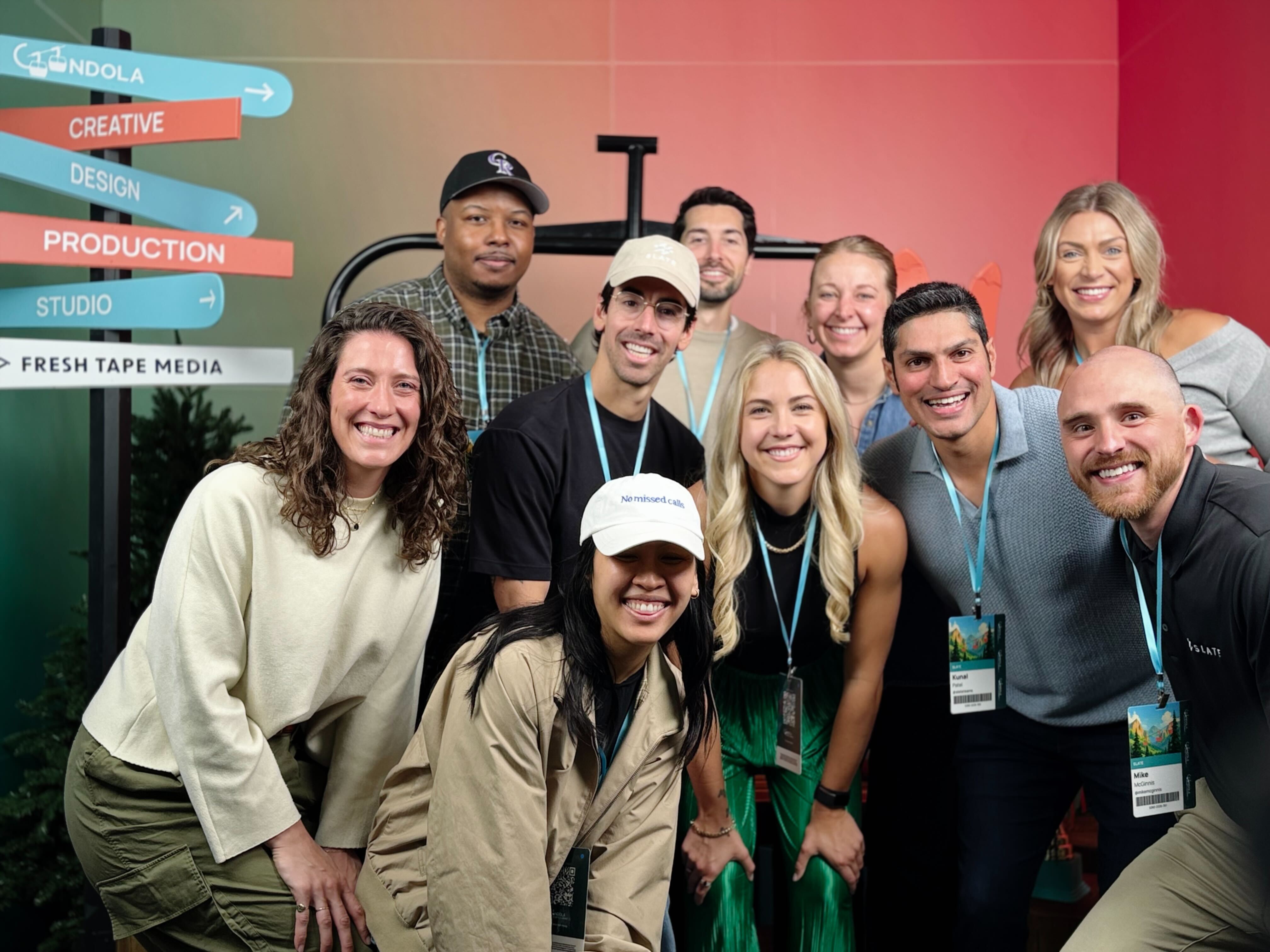 Group of smiling conference attendees posing together in front of a colorful Gondola event backdrop, with directional signs for creative, design, production, and studio visible on the left.
