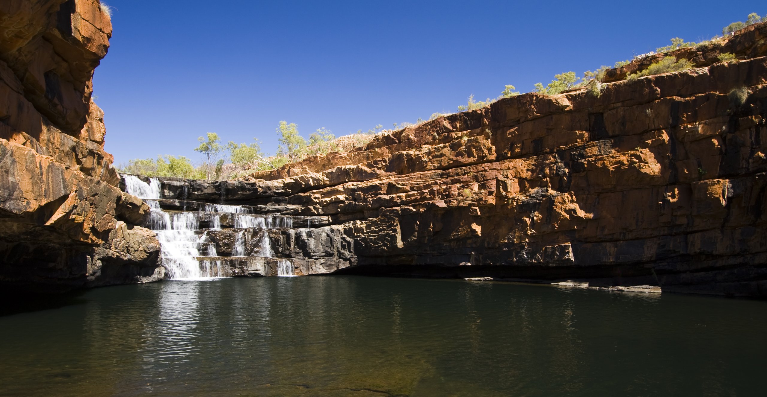 Bell Gorge in the Kimberley, Western Australia, a layered red-rock cascade flowing over horizontally-bedded sandstone into a clear plunge pool.
