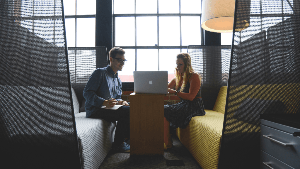 Two people sit facing each other in a small meeting area with high-backed seats on either side of a narrow table. A laptop is open on the table between them, and one person is writing in a notebook. Large windows fill the background, and a tall floor lamp is visible on the right.