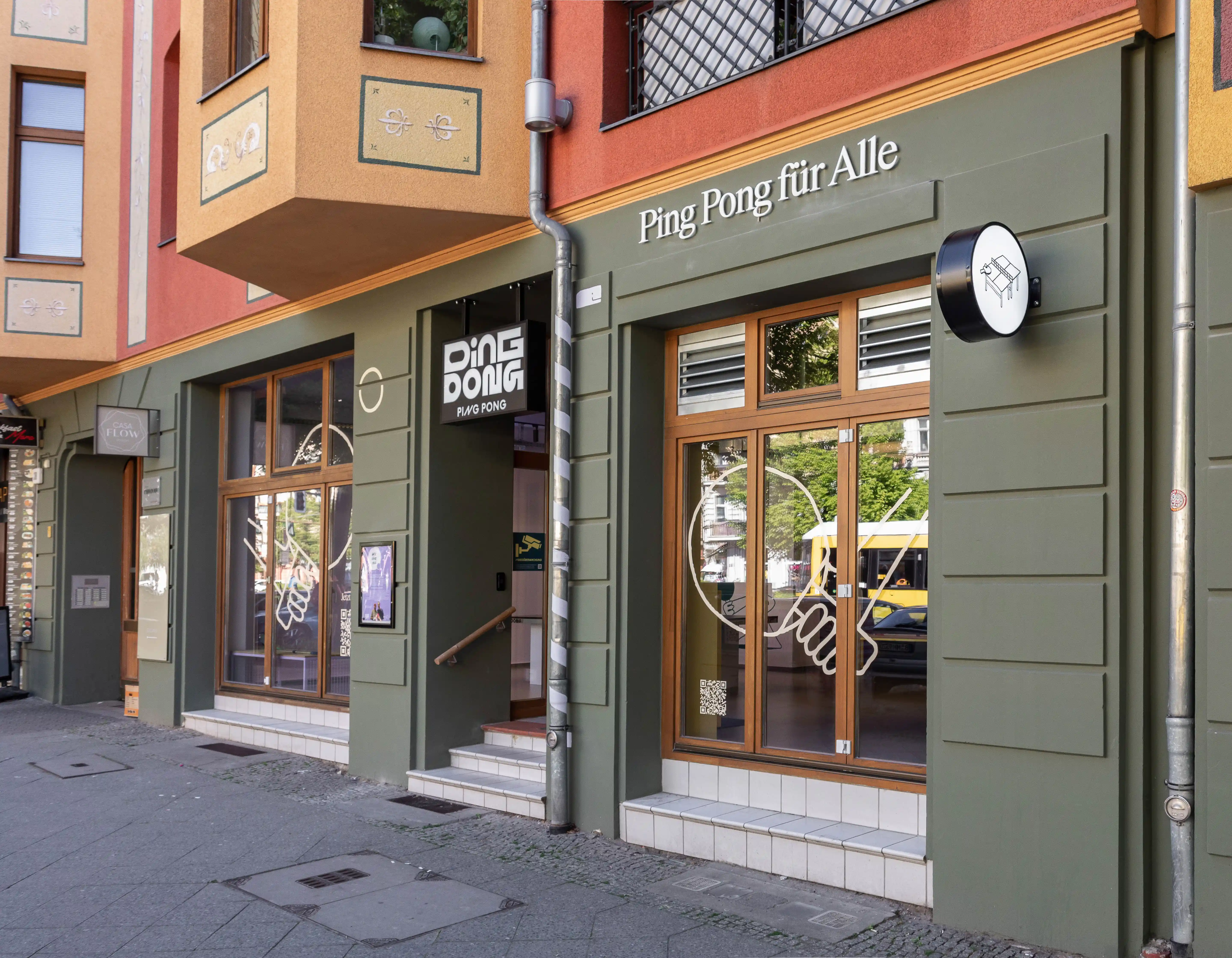 Street facade of DING DONG PING PONG in olive green with wooden window frames, Ping Pong für Alle lettering, a black logo sign and hand graphics on the shop windows.
