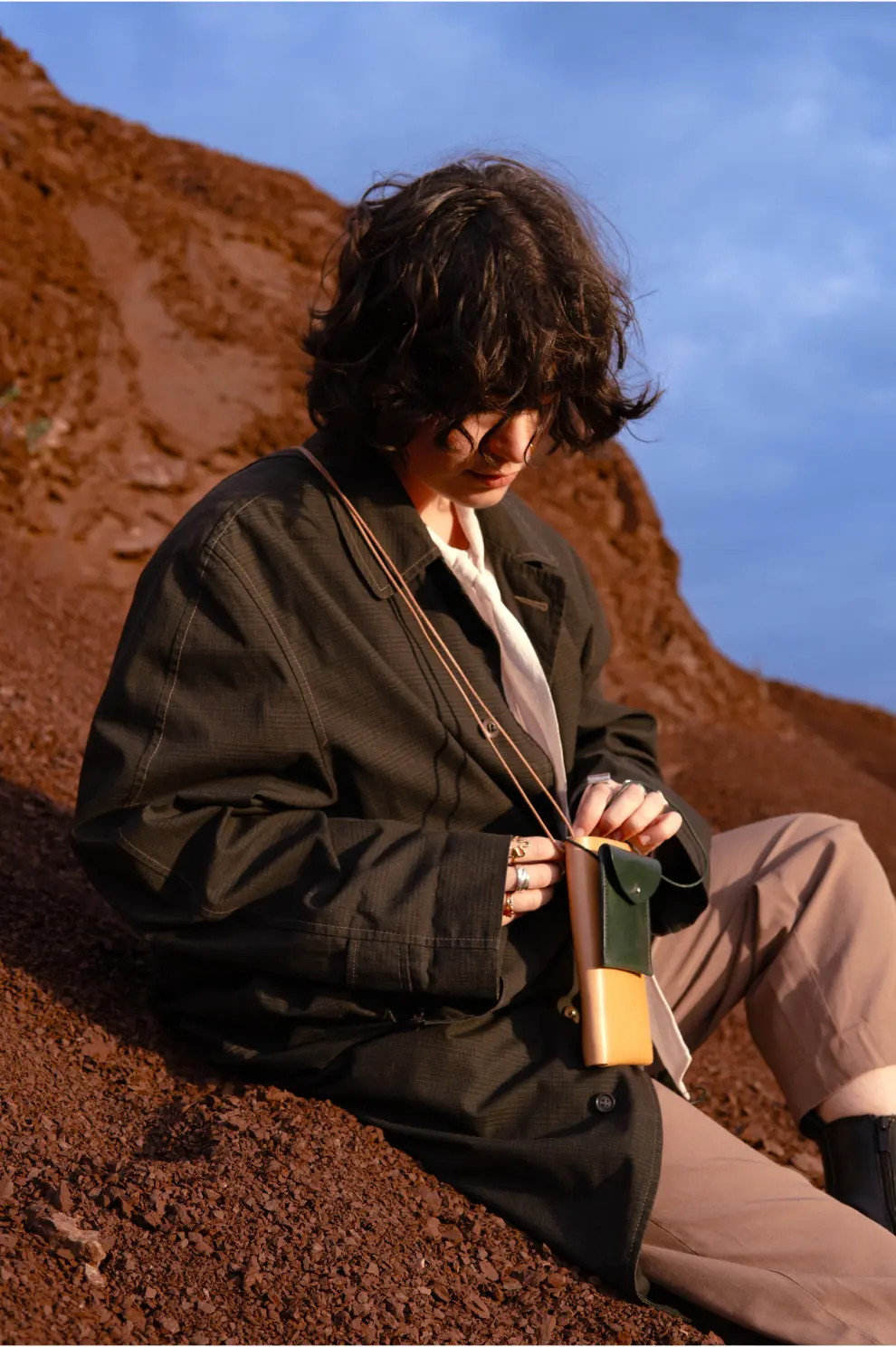 Anaïs Brancheriau sitting on red gravel, looking down at a small crossbody pouch, lit by warm late-day sunlight.
