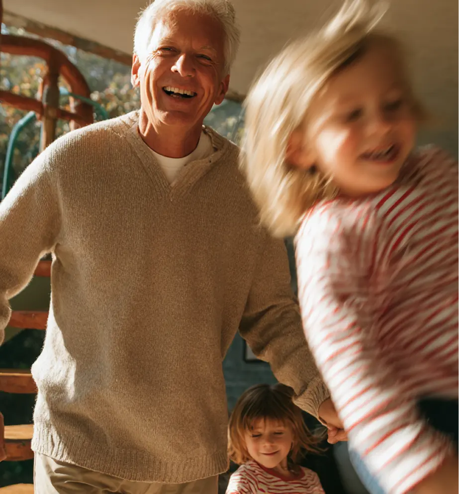 Laughing older man playing with children indoors, holding hands and moving together, with motion blur capturing a joyful, energetic moment in warm light.