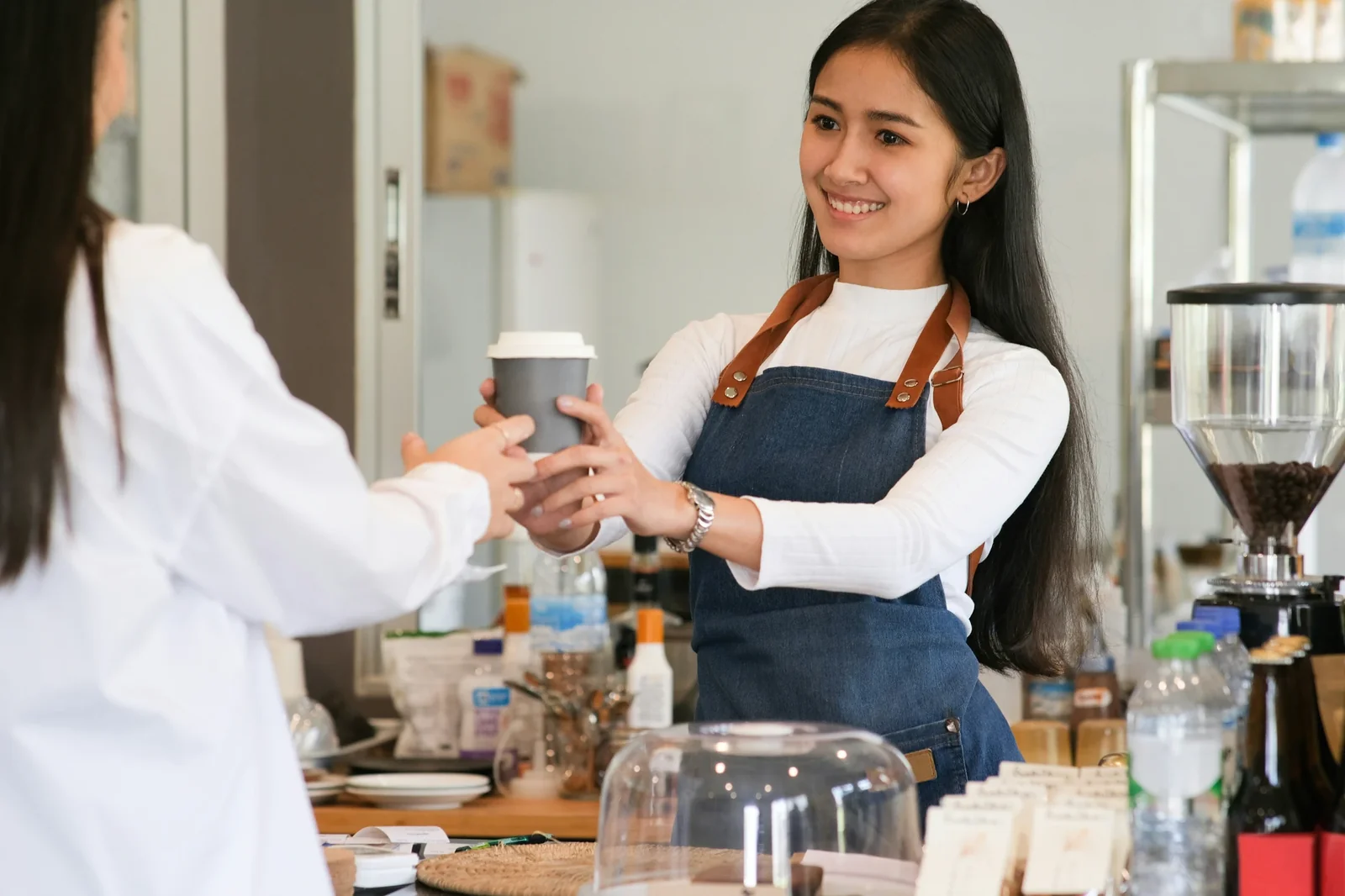 Smiling barista in denim apron hands coffee cup to customer in modern café setting.