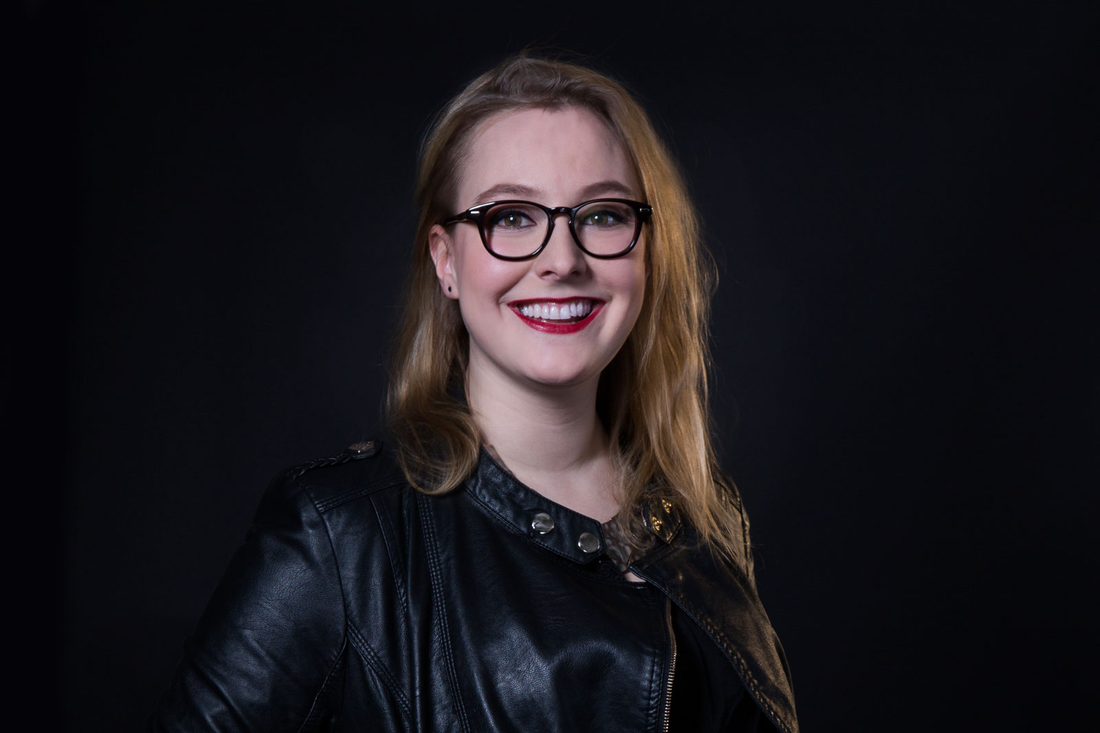 A woman with long blonde hair and round glasses wears a black leather jacket. She is smiling and looking directly at the camera against a black background.