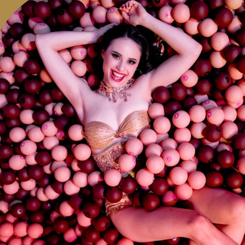 A woman in a gold outfit is lying in a pool filled with pink and brown spherical objects, smiling at the camera.