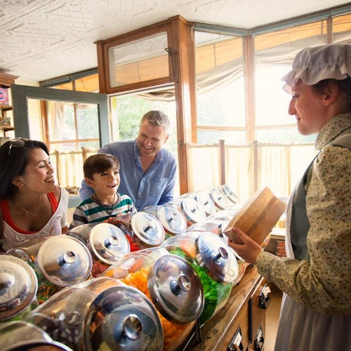 A family smiles at a shopkeeper in a bonnet behind a counter filled with large jars of colorful candy.
