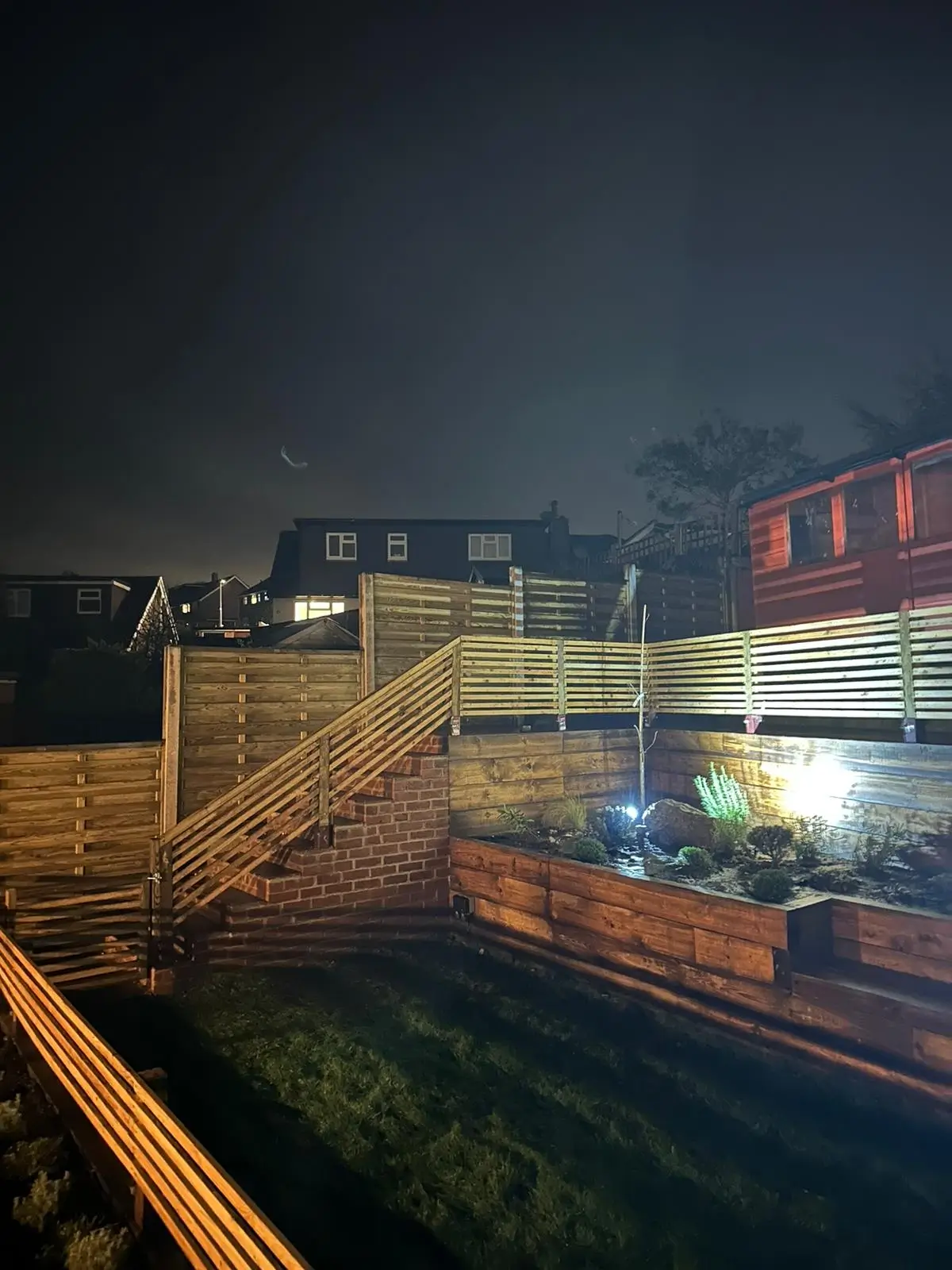 A dimly lit urban scene at night, showing buildings, a fence, and scattered debris near a body of water.