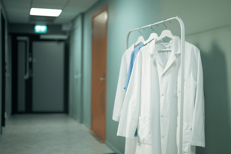 En gang i et fastlegekontor med en hvit legefrakk hengende på et stativ og dører i bakgrunnen.
