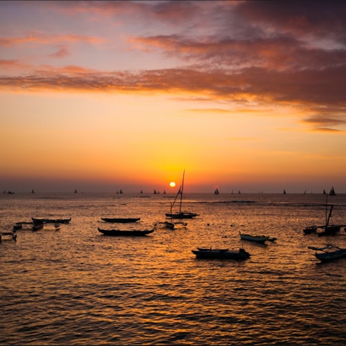 เรือใบและเรือพายลอยอยู่บนทะเลที่สงบในยามพระอาทิตย์ตกดิน โดยมีดวงอาทิตย์ใกล้ขอบฟ้าและท้องฟ้าถูกระบายด้วยเมฆสีส้มและสีม่วง
