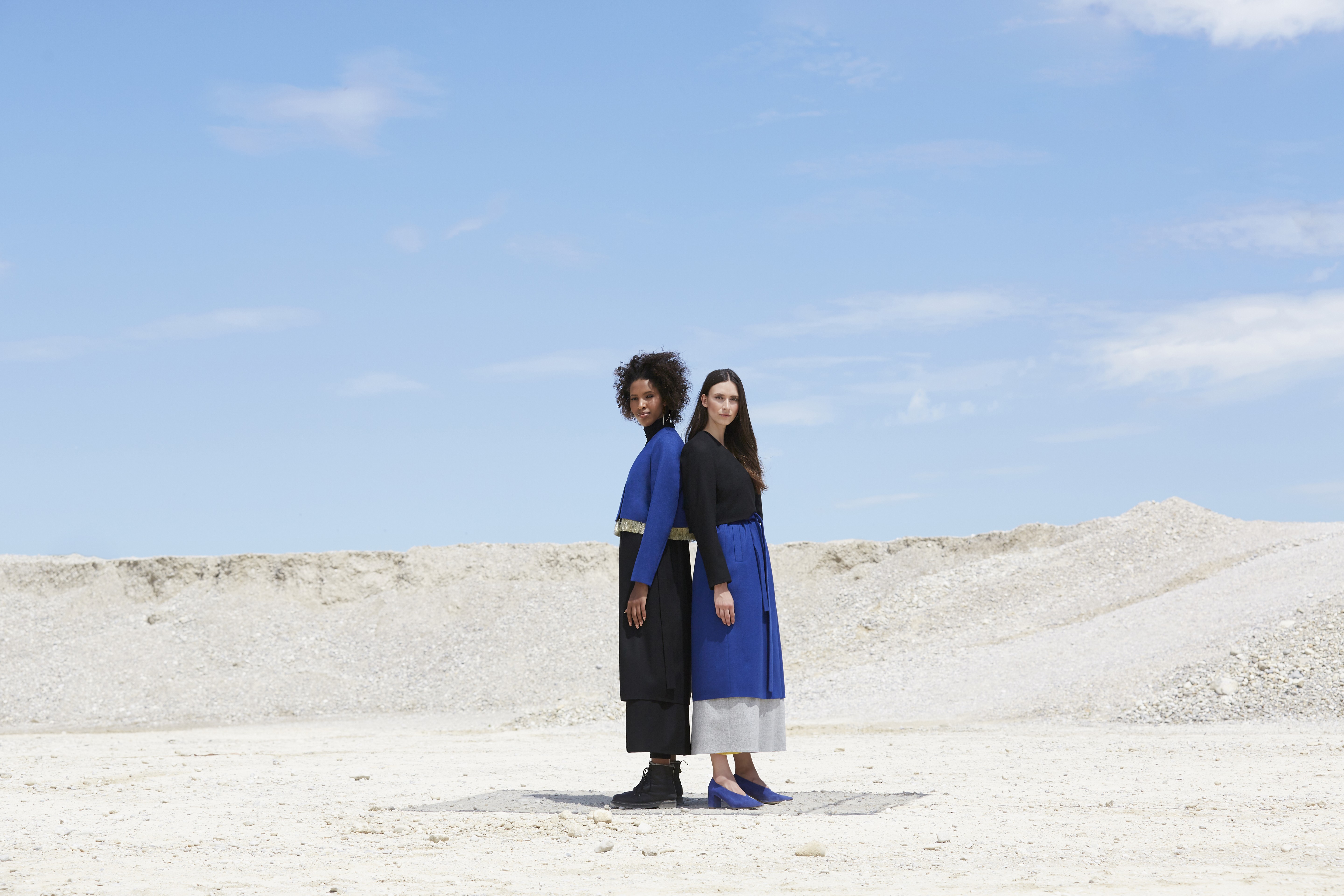 Two individuals stand back-to-back in a barren, sandy landscape under a clear blue sky, both wearing stylish black and blue outfits that contrast with the stark surroundings.