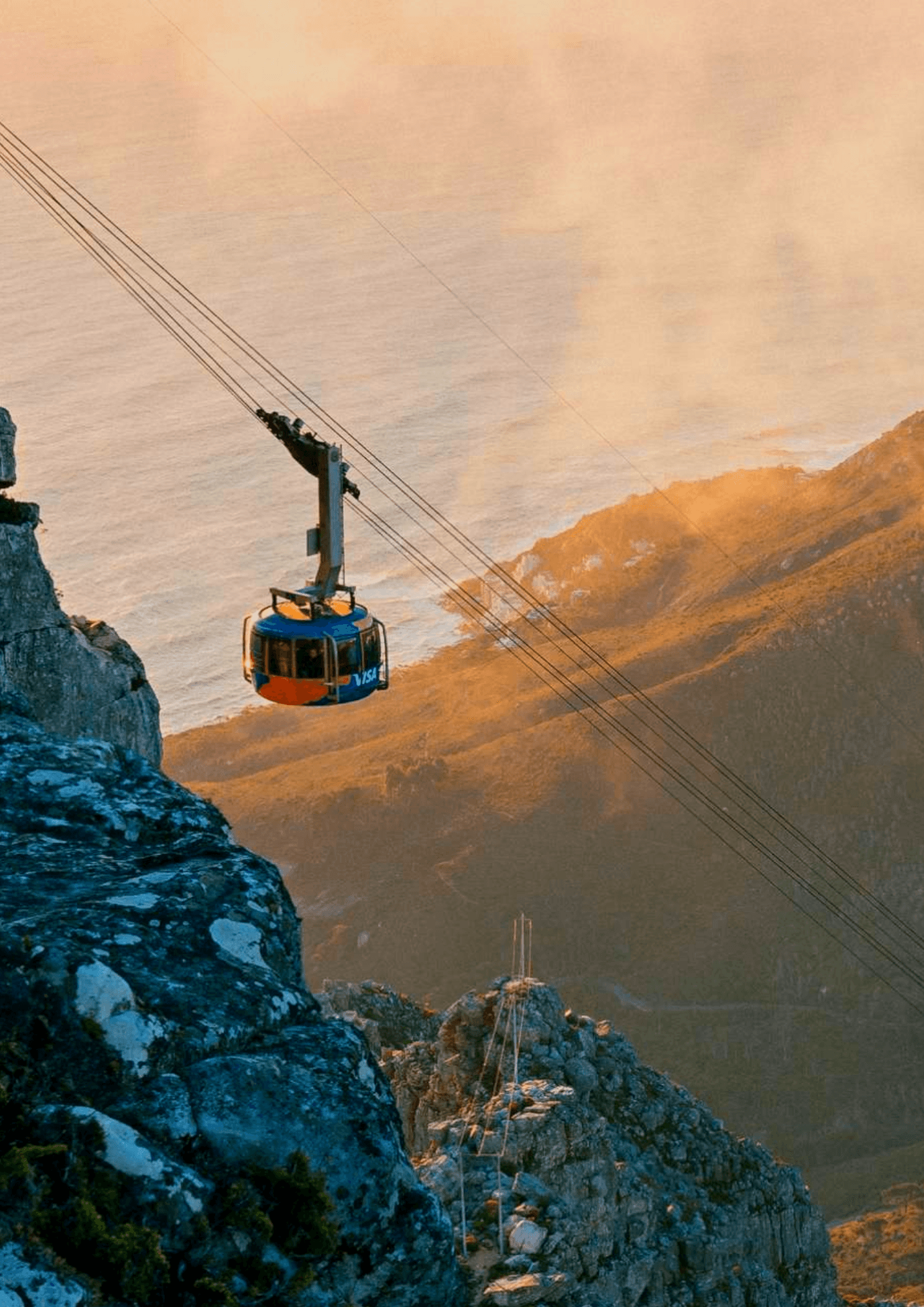 A cable car ascends a steep mountainside with a breathtaking coastal view in the background, under a soft, golden sunset sky.