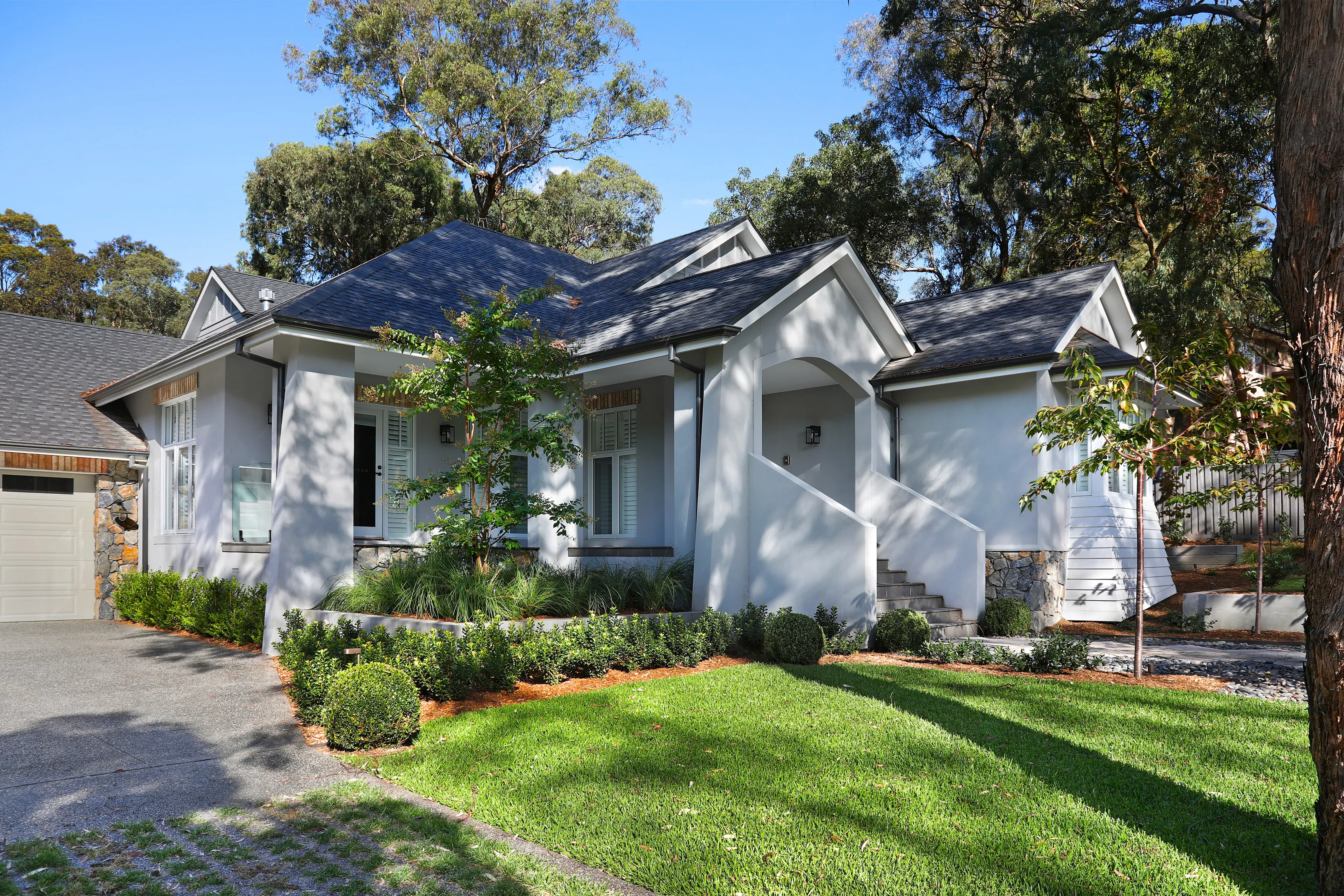 Front of the Montmorency Residence with a rendered facade, stacked-stone base, multiple gabled grey roofs, plantation shutters, an arched portico and clipped hedges along the driveway