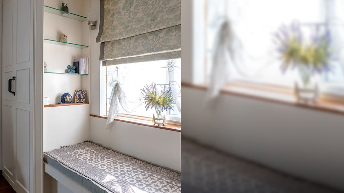 A white window seat with half-drawn-up grey patterned blinds and shelves with ornaments. The ornaments include small ornamental owls, plants, and a photo frame.